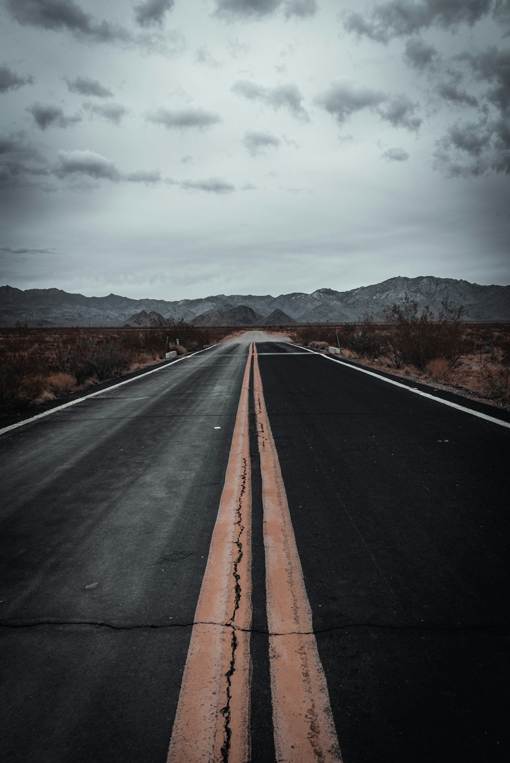 A long, straight road in a desert landscape with mountains in the background and a cloudy sky overhead.