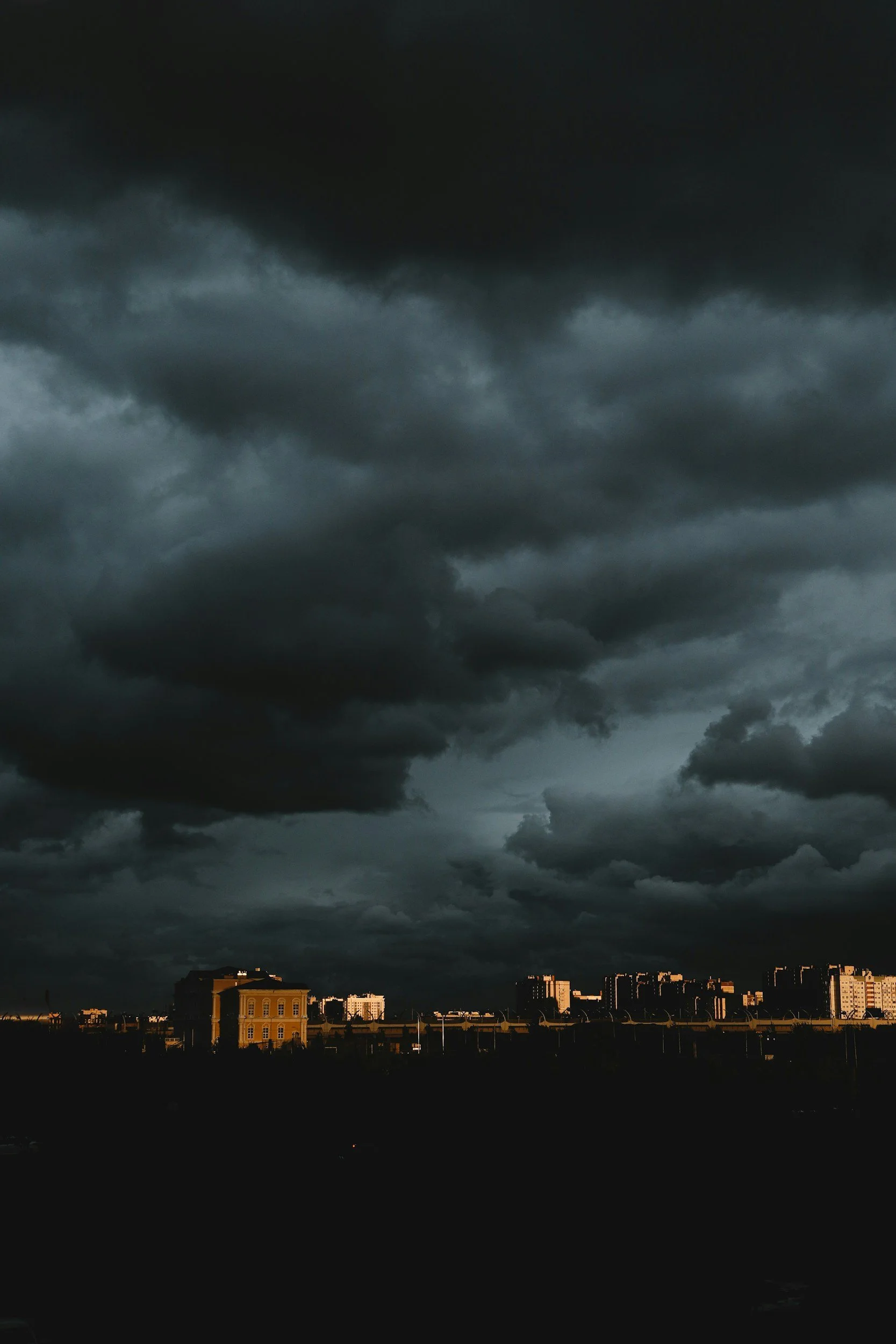 Dark, stormy clouds over a cityscape with a mix of older and modern buildings, illuminated by a faint light from the setting or rising sun.