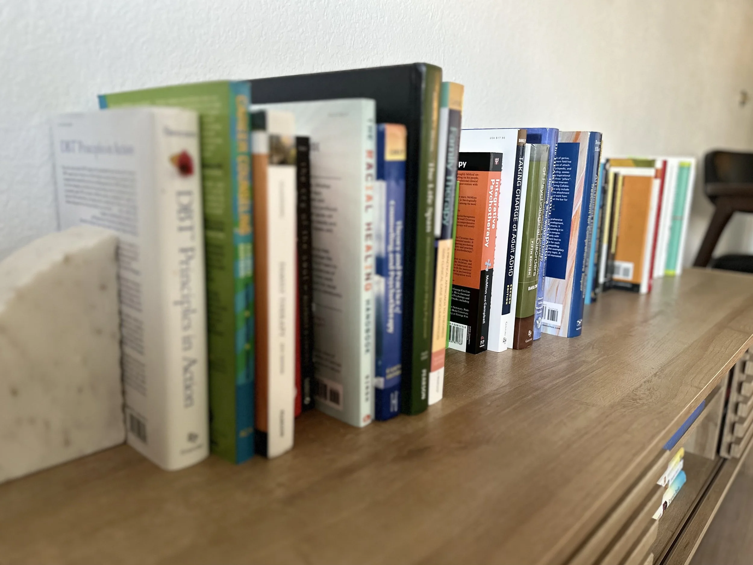 A row of books on a wooden table, with some books stacked vertically and others leaning; the books are of various colors and sizes.