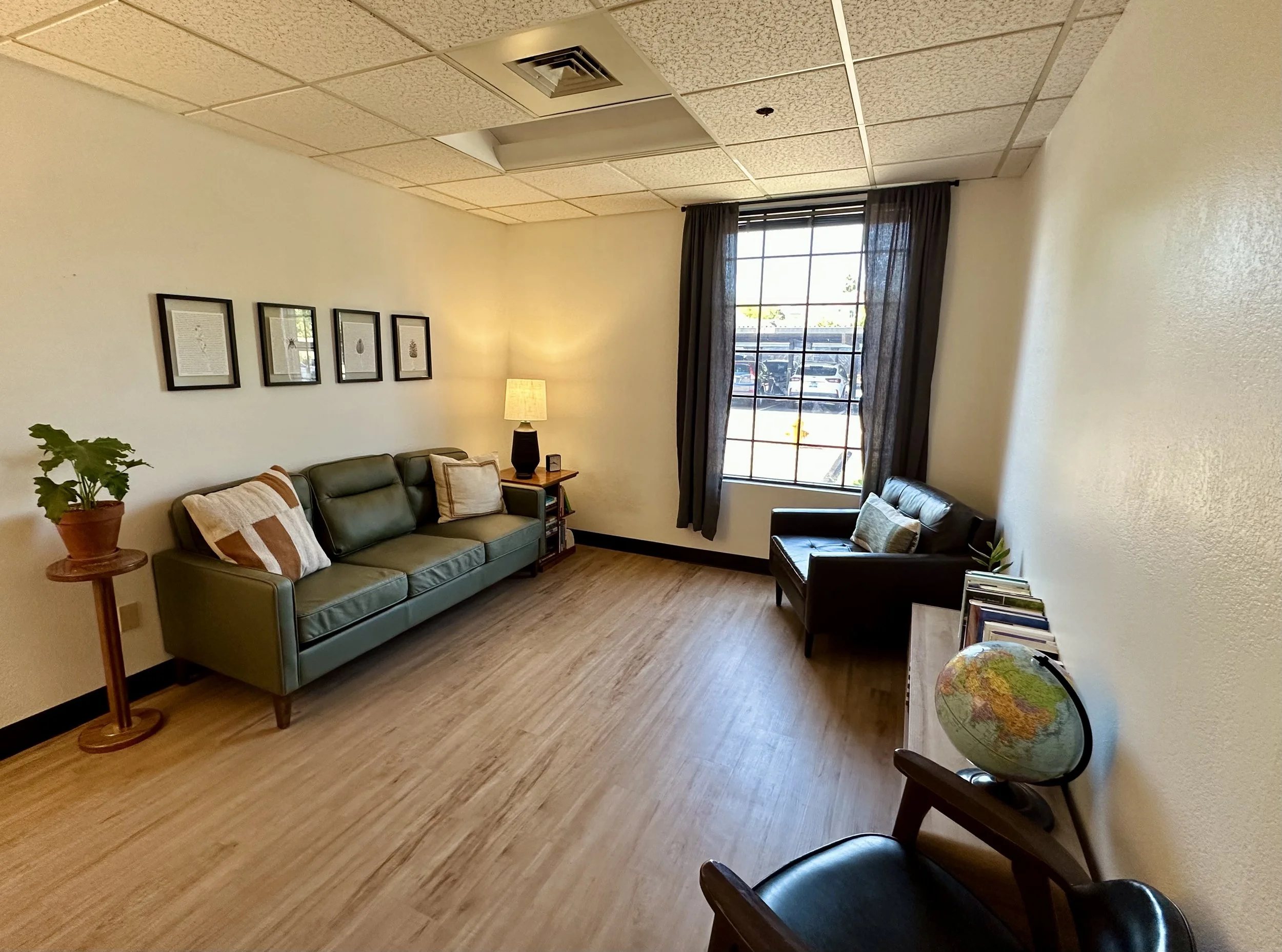 Simple office or waiting room with a green sofa, a black armchair, a wooden side table with a globe and books, a window with black curtains, framed artwork on the wall, a small side table with a lamp, and a few pillows.