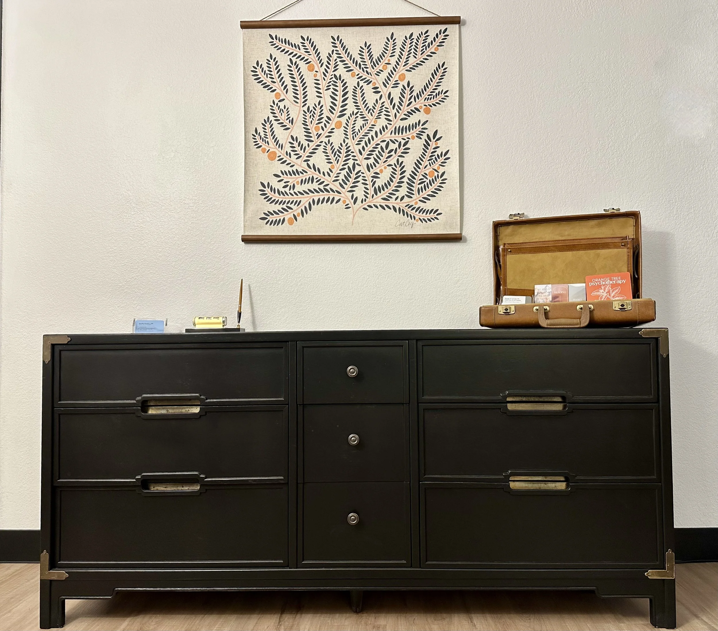 Black dresser with multiple drawers, topped with a small open suitcase displaying various items, against a plain wall with a decorative leaf-patterned wall hanging above.