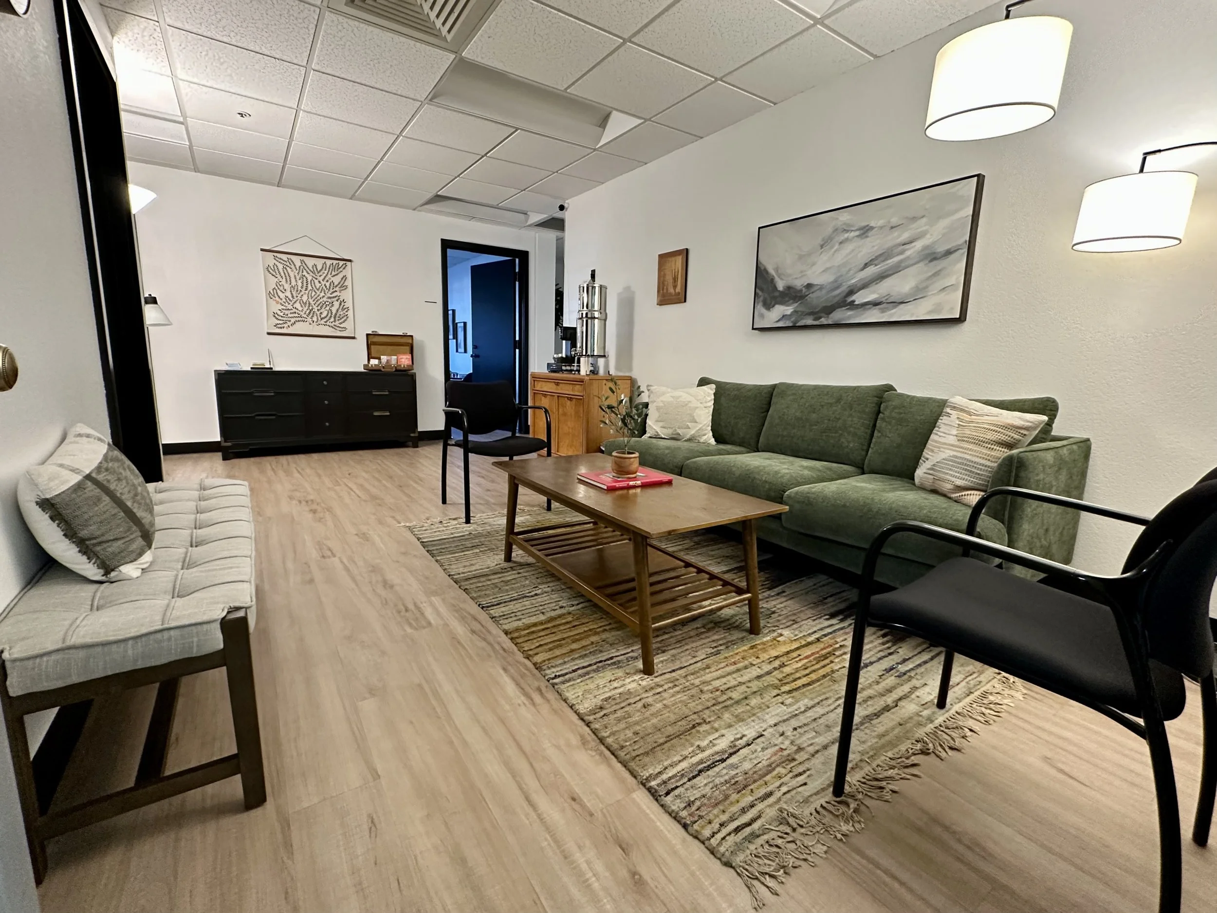 Modern waiting room with a green sofa, two armchairs, a wooden coffee table on a rug, a low bench, and wall art on white walls.