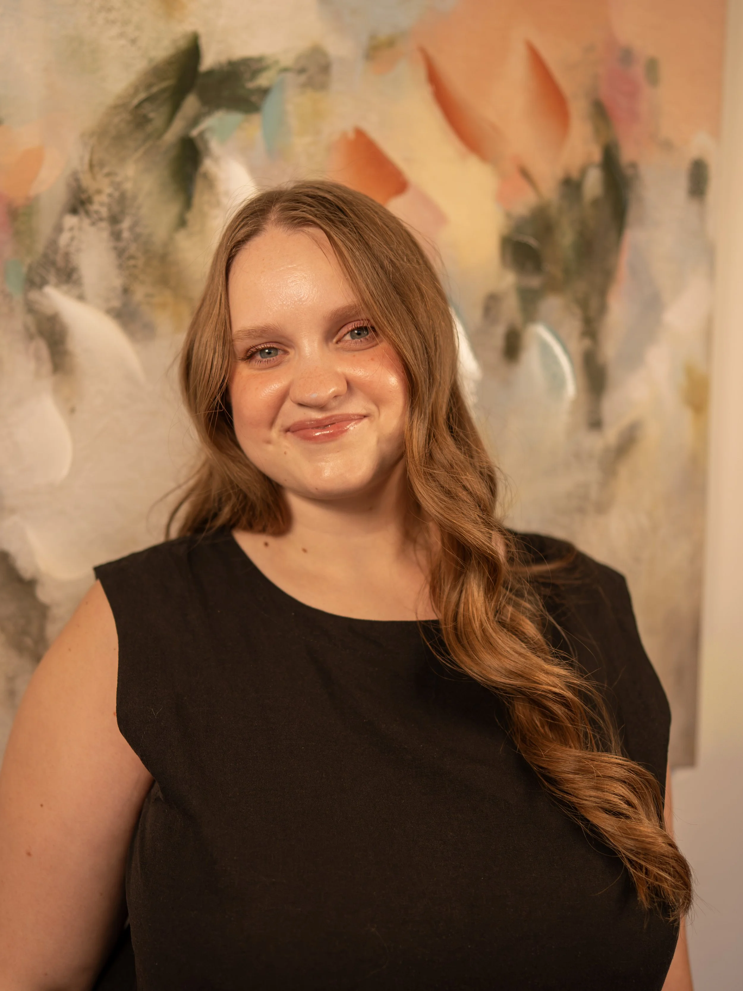 A woman with long wavy red hair and a black sleeveless top smiling in front of an abstract painting with warm colors and leaf-like shapes.