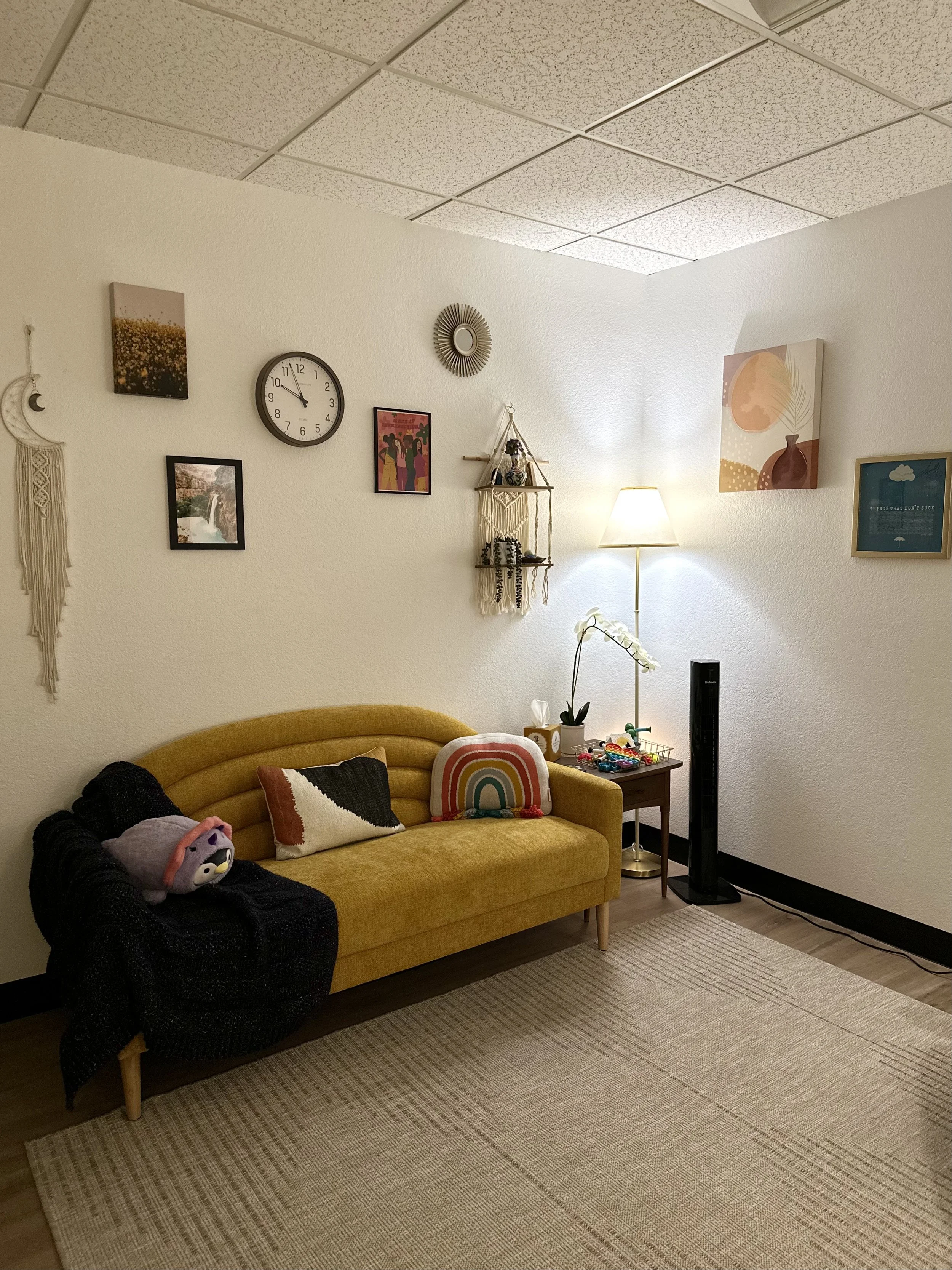 A mustard yellow sofa with three decorative pillows, including a rainbow cushion, in a cozy living room. A small wooden side table next to it holds a white orchid, tissues, and toys. A tall standing fan is nearby, and a floor lamp provides warm lighting. The wall above has a clock, artwork, and decorative wall hangings, creating a warm and inviting atmosphere.