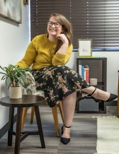 Woman sitting on a chair with a floral skirt and yellow top, smiling with glasses, in a cozy room with window blinds and a small table with plants and books.