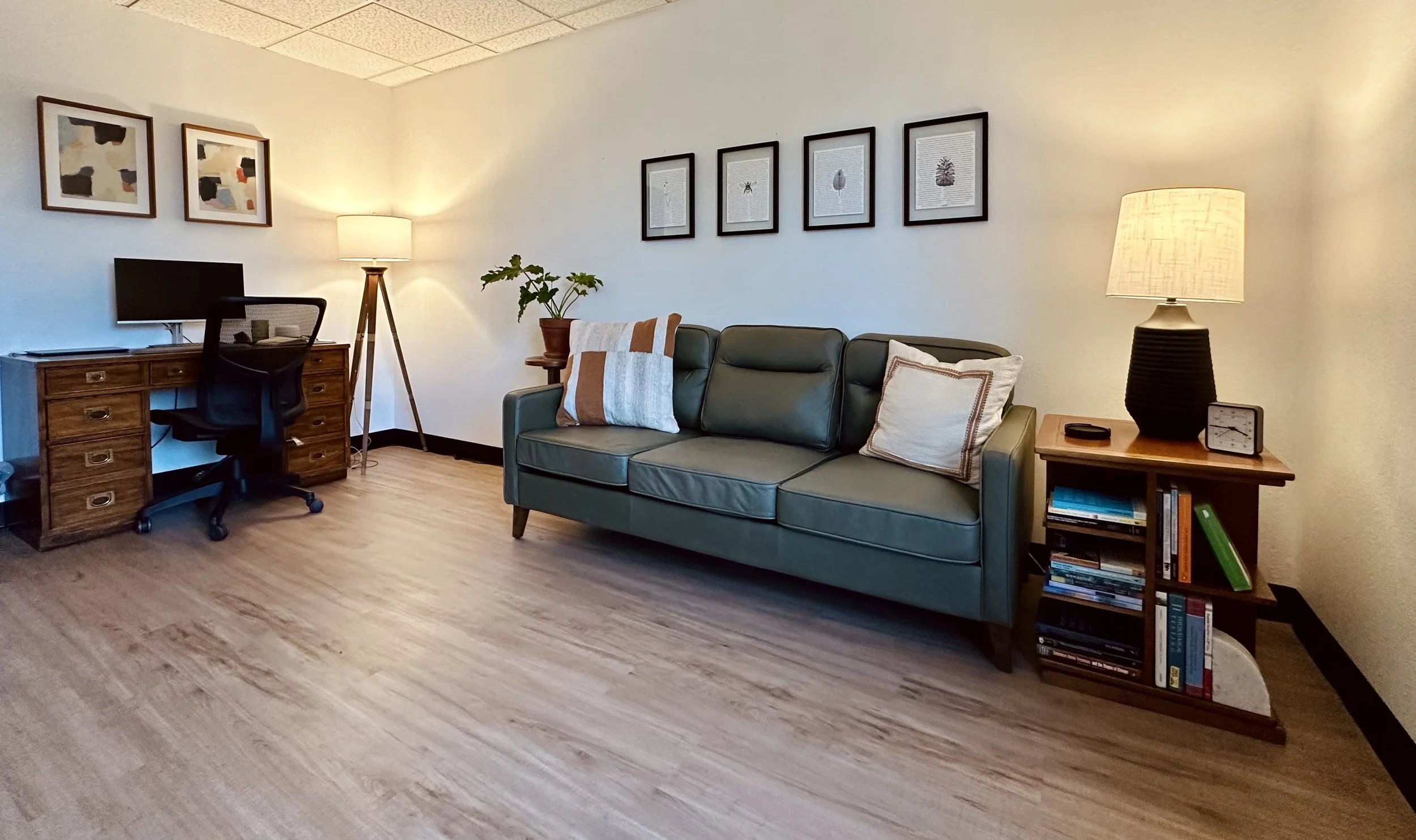 A living room with a desk and chair on the left, a green sofa with pillows in the center, a bookshelf with books and a table lamp on the right, and framed artwork on the wall.