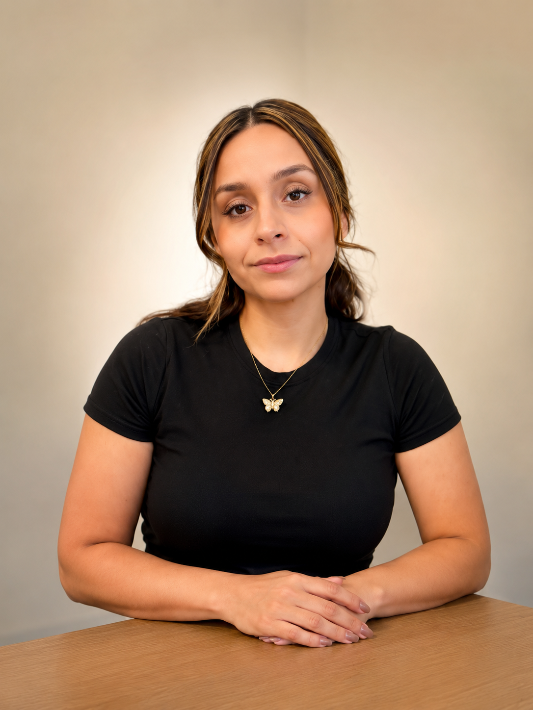 A young woman with light skin and brown hair sitting at a wooden table, wearing a black T-shirt and a butterfly necklace, looking at the camera with a subtle smile.