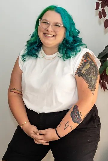 Woman with blue and green hair, wearing glasses and a white t-shirt, smiling, with tattoos on her arms, standing against a neutral background with a plant.