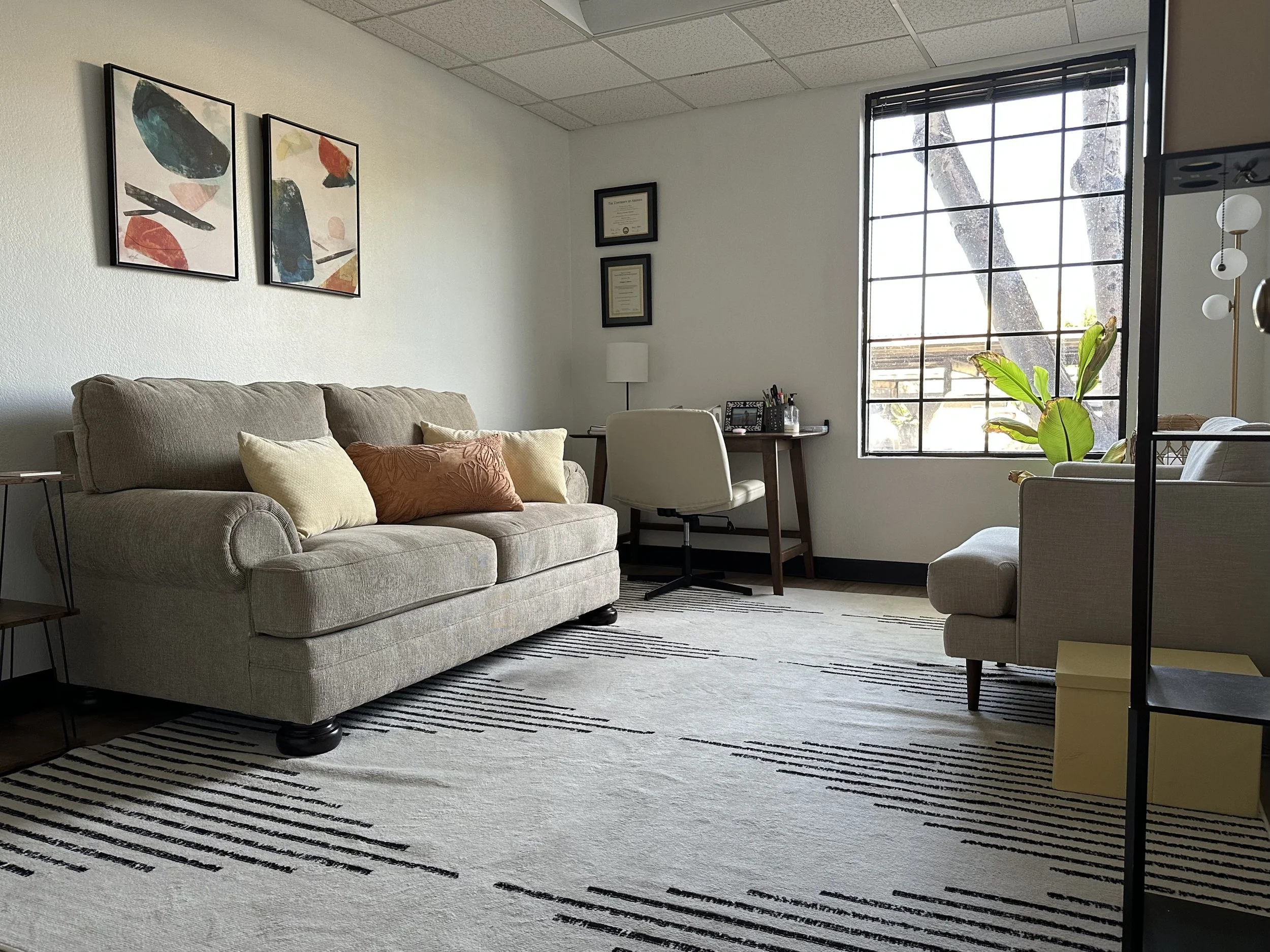 Modern living room with a beige sofa, abstract wall art, desk with chair, large window, and indoor plant.