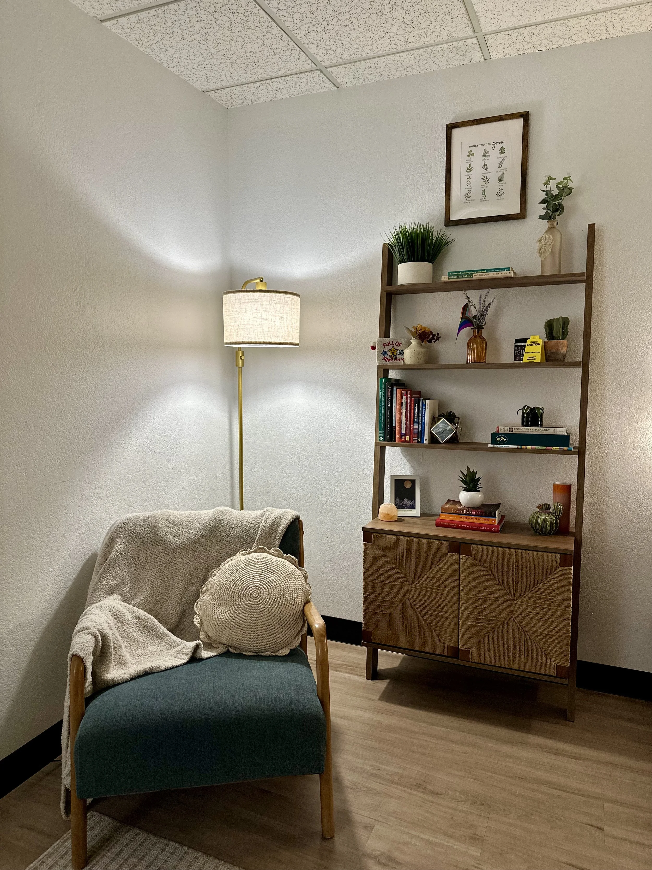 A cozy corner with a teal armchair draped with a beige blanket and a round crochet pillow. Behind the chair is a tall floor lamp with a beige shade. To the right, a tall wooden bookshelf holds plants, books, and decorative items. The room has off-white walls, a ceiling with acoustic tiles, and light wood flooring.
