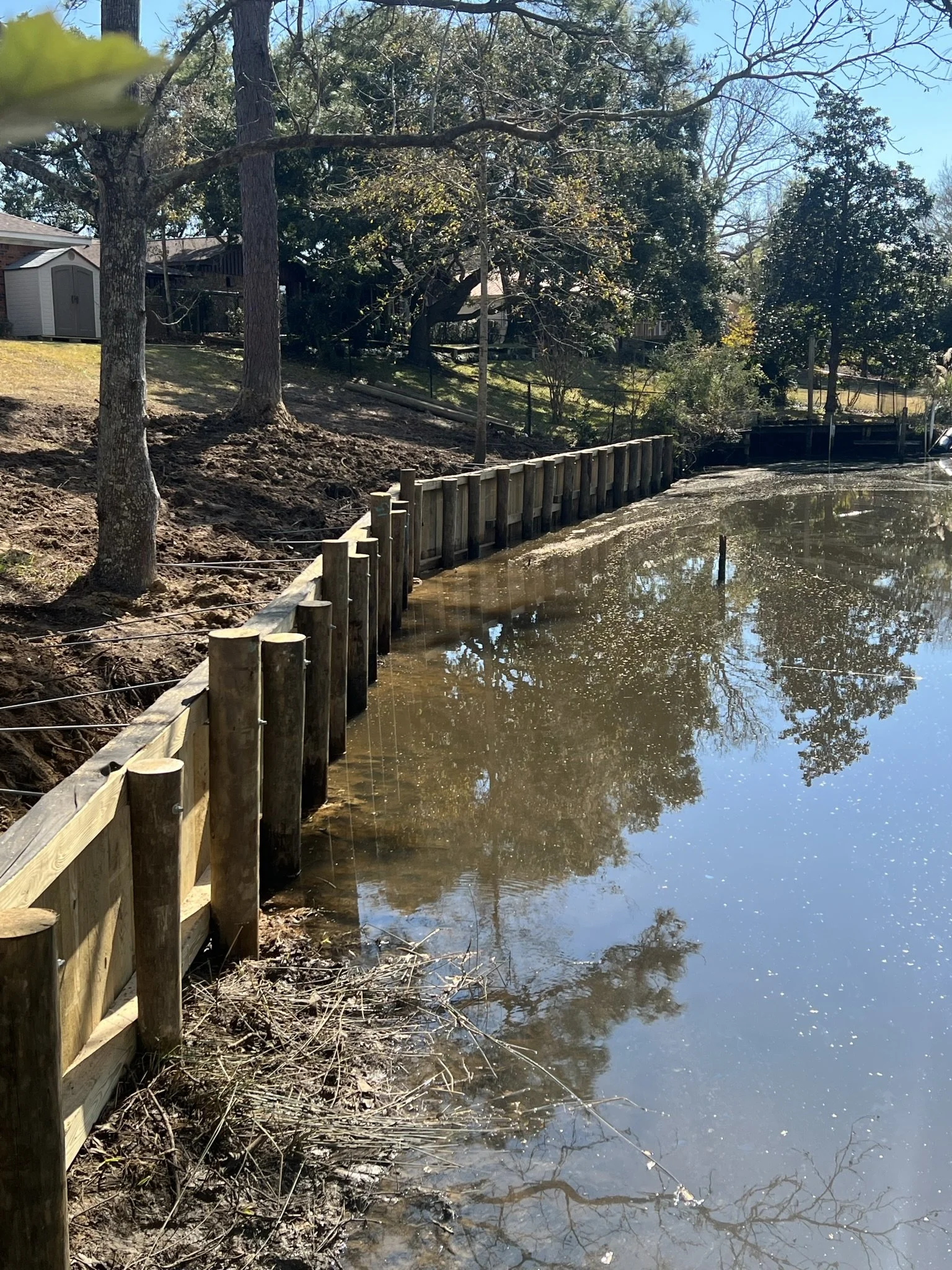 Wooden barrier along the edge of a muddy riverbank with trees and a house in the background, under a clear blue sky.