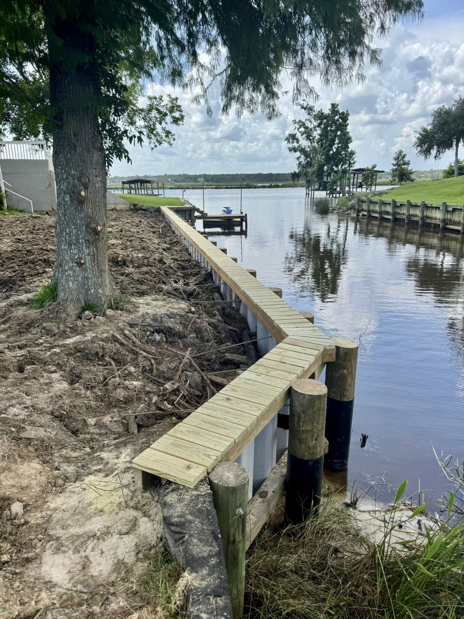A newly built wooden dock along a riverbank, with trees and green grass in the background under a partly cloudy sky.