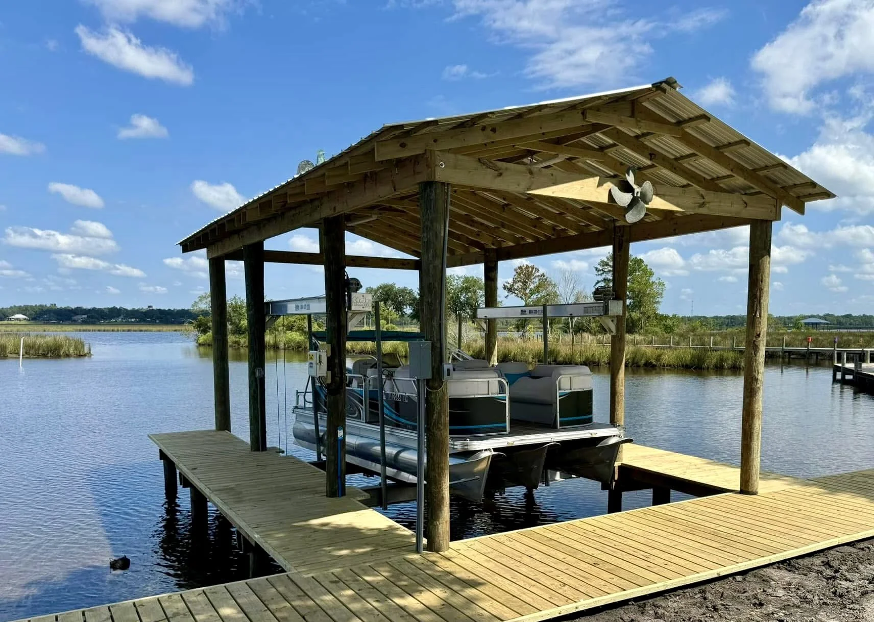 A boat dock with a covered shelter over a pontoon boat on a river, with a wooden walkway leading to it and open water in the background under a partly cloudy sky.