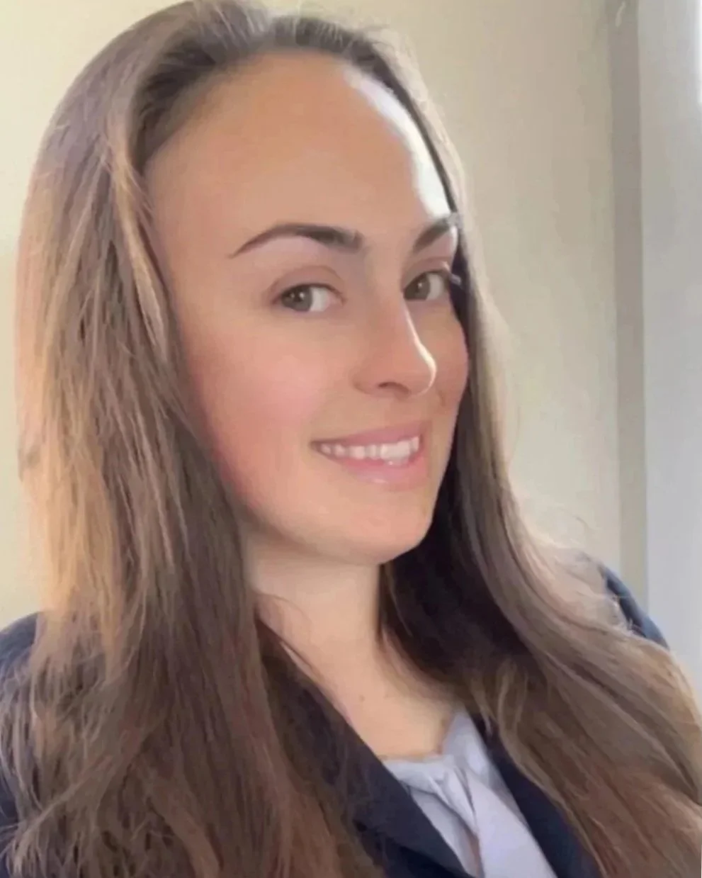 A young woman with long, wavy light brown hair, smiling, wearing a dark jacket over a light-colored blouse, standing inside near a window.