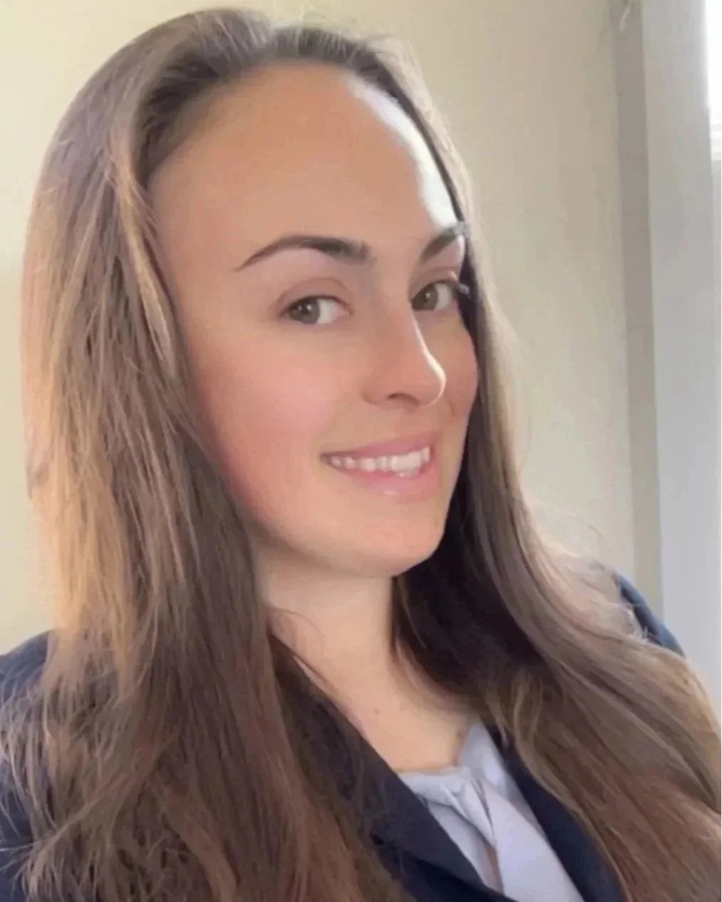A young woman with long, wavy light brown hair, smiling, wearing a dark jacket over a light-colored blouse, standing inside near a window.