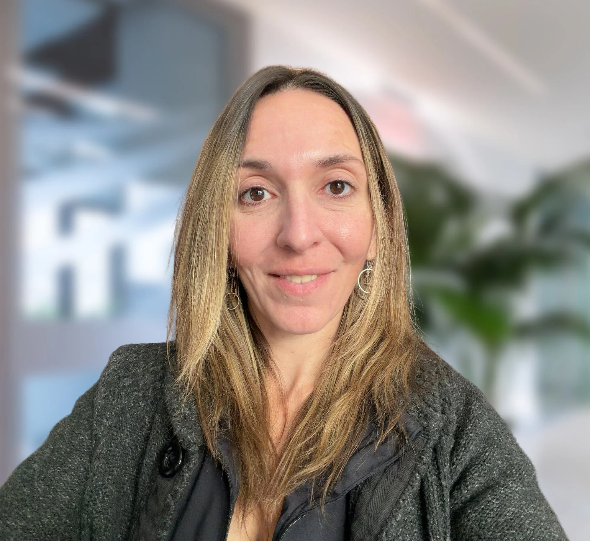 Close-up of a woman with curly brown hair, wearing a gray sweater, earrings, and a delicate necklace, sitting inside a vehicle.