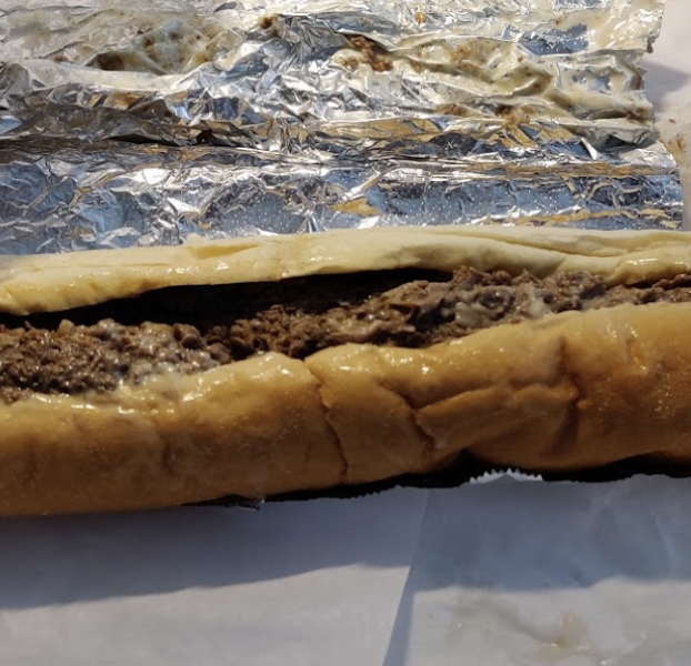 A close-up of a cheesesteak sandwich with a long bun, ground beef filling, and aluminum foil in the background.