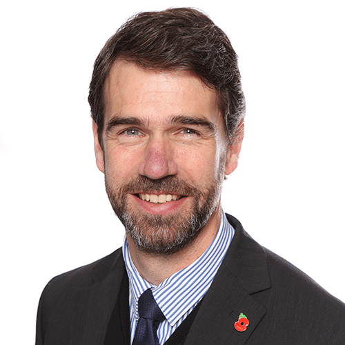 Ben Riggott. Headshot of a man with dark hair and a beard, wearing a suit and tie, smiling against a white background.