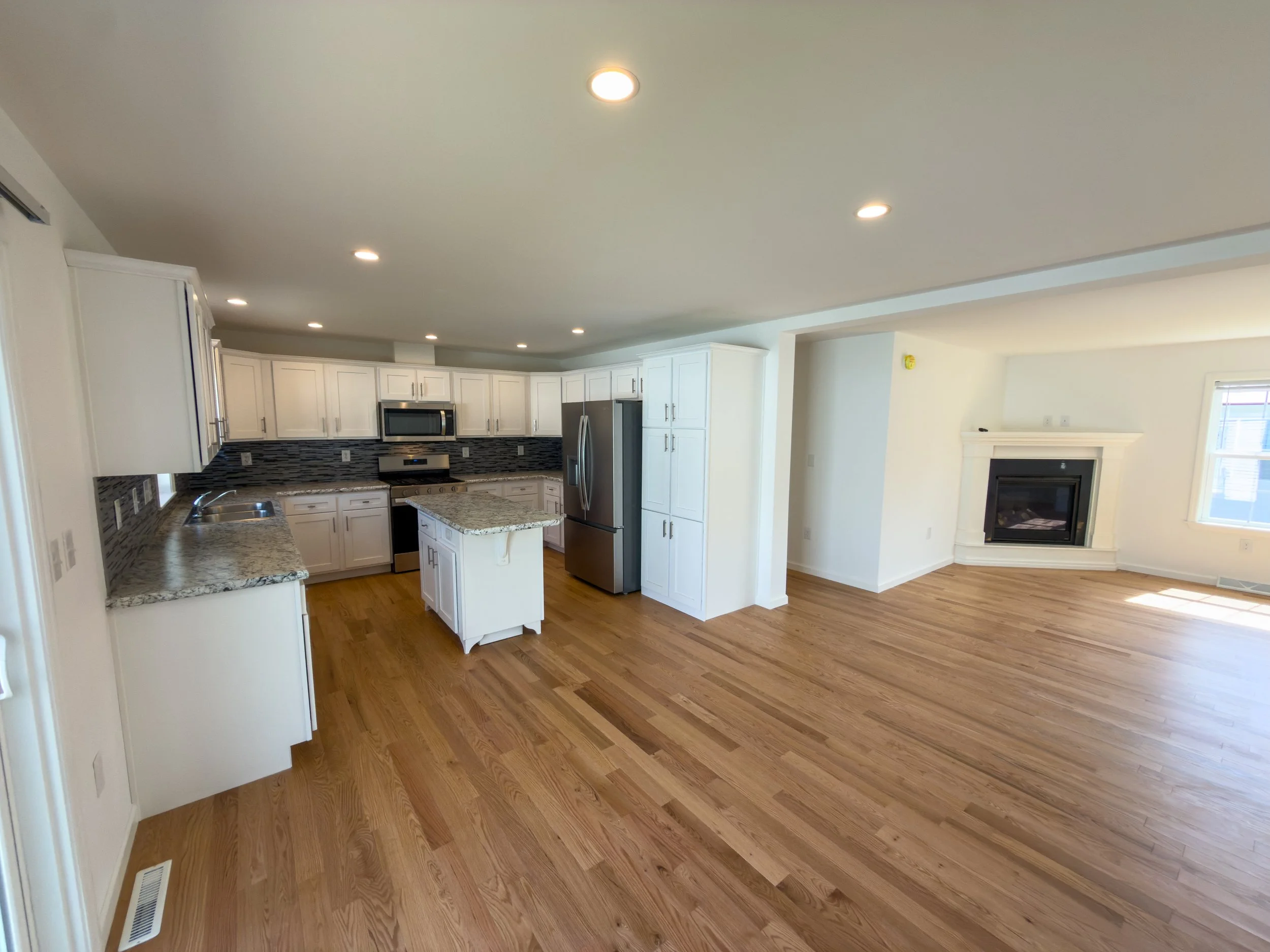 Modern open kitchen with white cabinets, countertops, a central island, stainless steel appliances, hardwood floors, and a fireplace visible in the adjacent living room