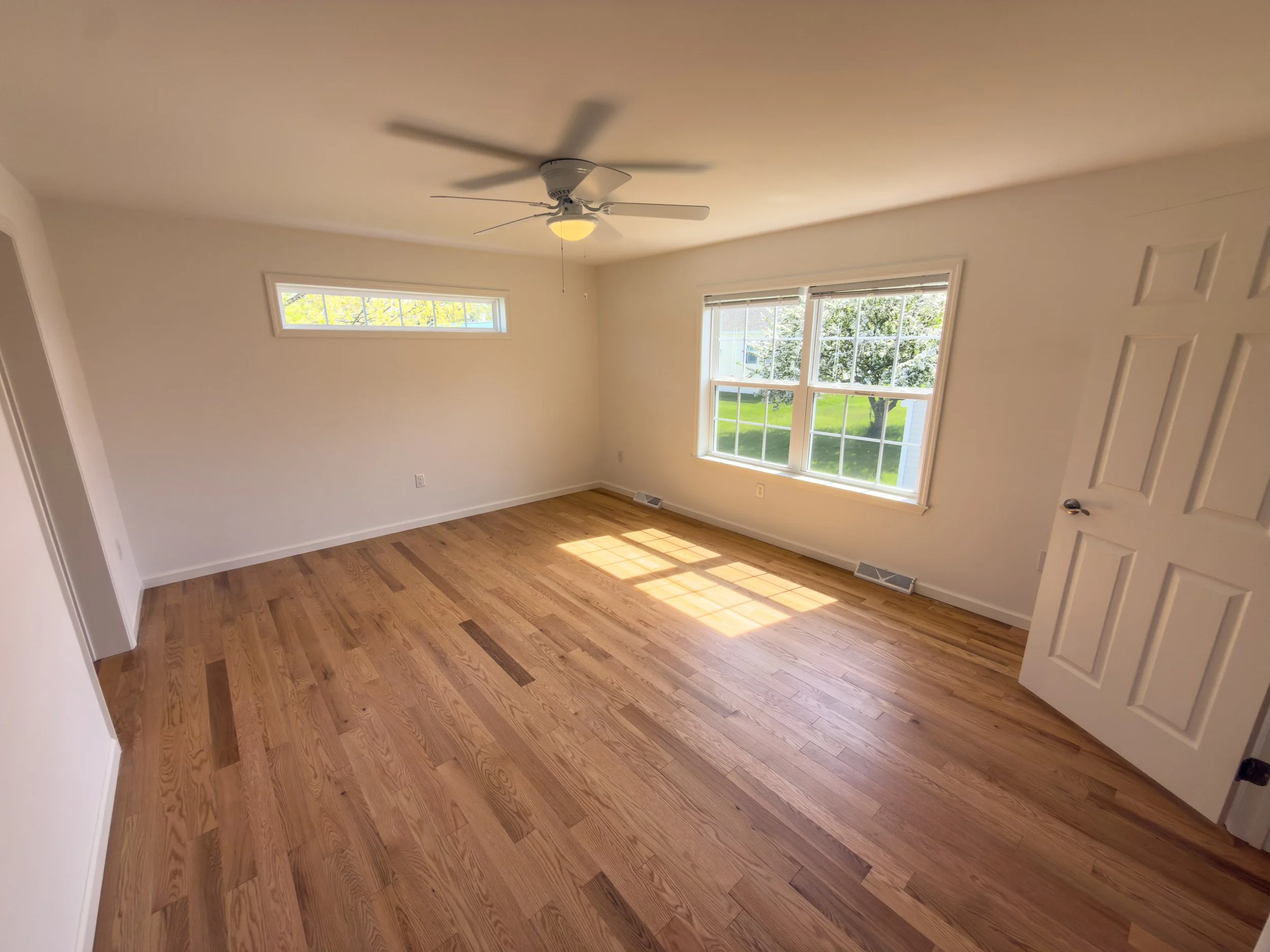 Bright and airy bedroom with gleaming hardwood floors, ceiling fan, and dual windows overlooking lush greenery — a peaceful, light-filled bedroom in a new manufactured home