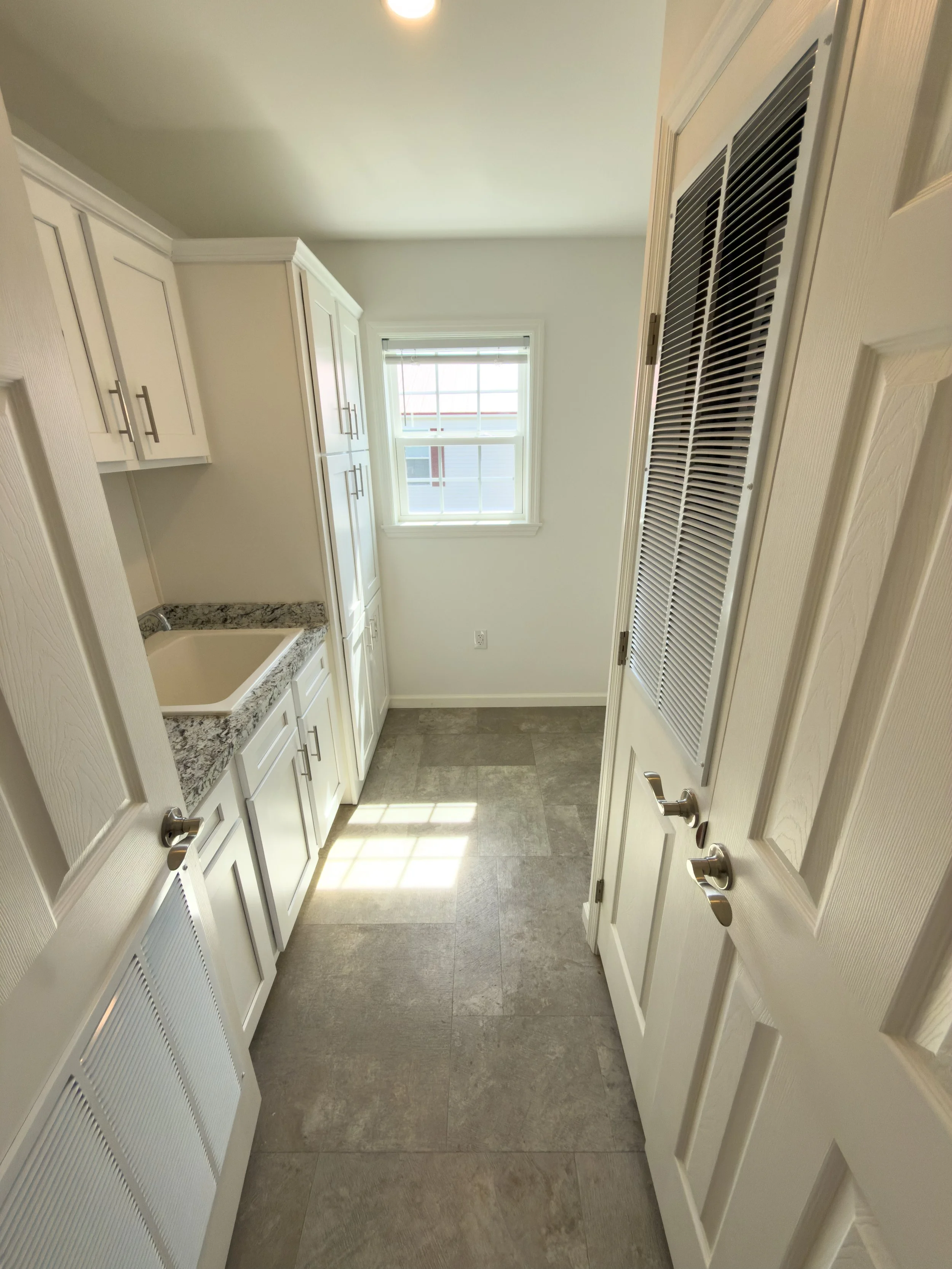Functional laundry or mudroom area with white cabinetry, countertop, a window with blinds, and tile flooring — convenient utility space at State Manufactured Homes