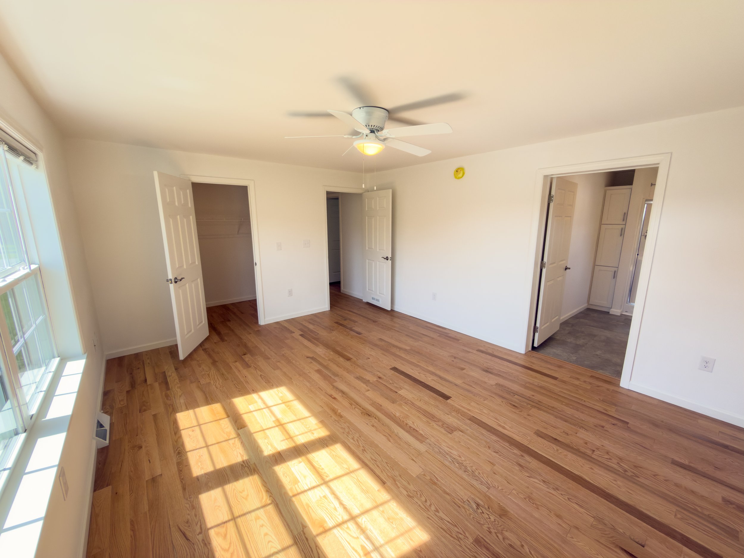 Generous master bedroom with warm hardwood floors, ceiling fan with light, three closet doors for ample storage, and sun-drenched flooring from bright windows