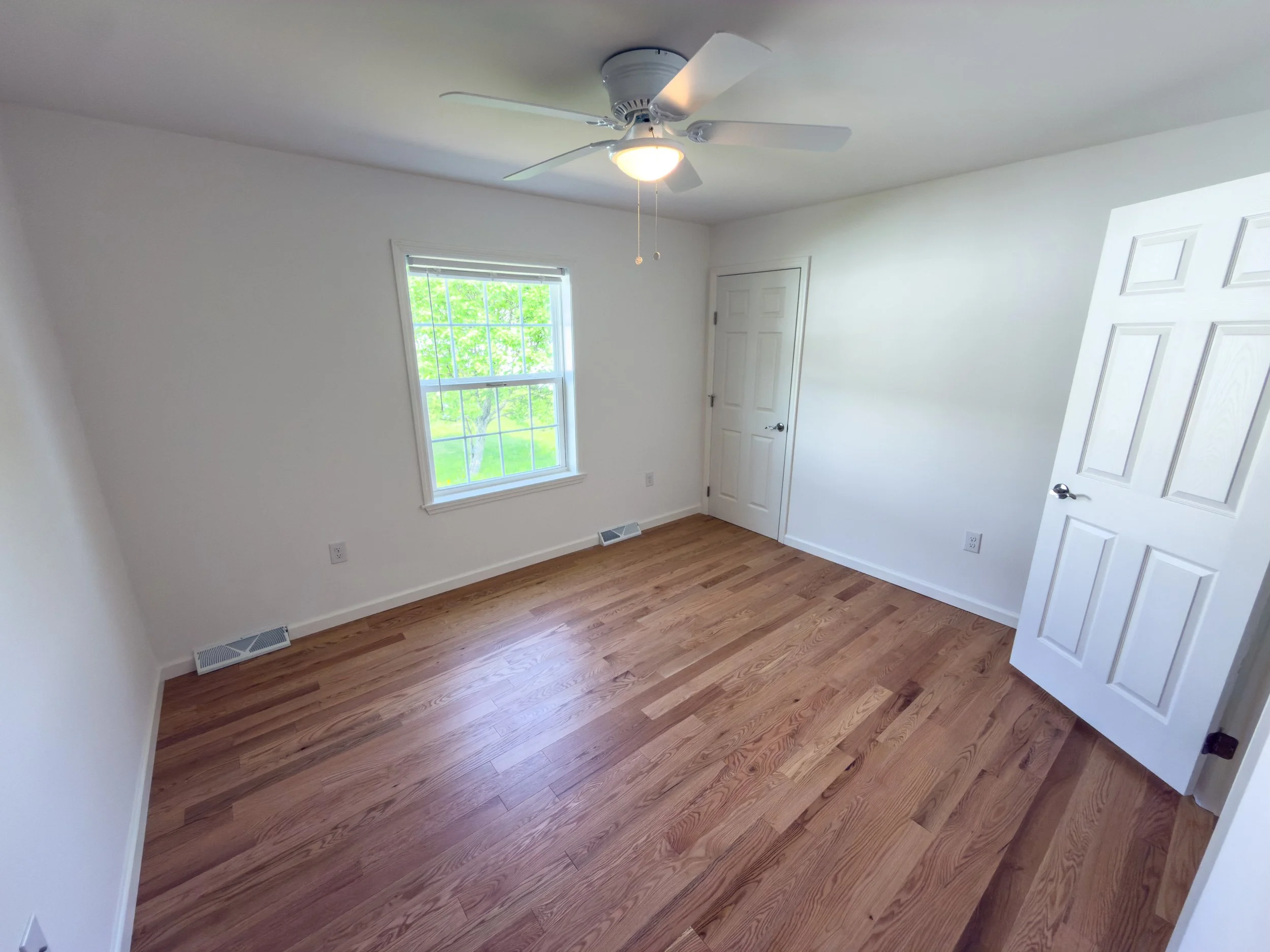 Cozy bedroom with hardwood floors, white walls, a ceiling fan with light, a window showcasing a serene view of green trees, and a white closet door