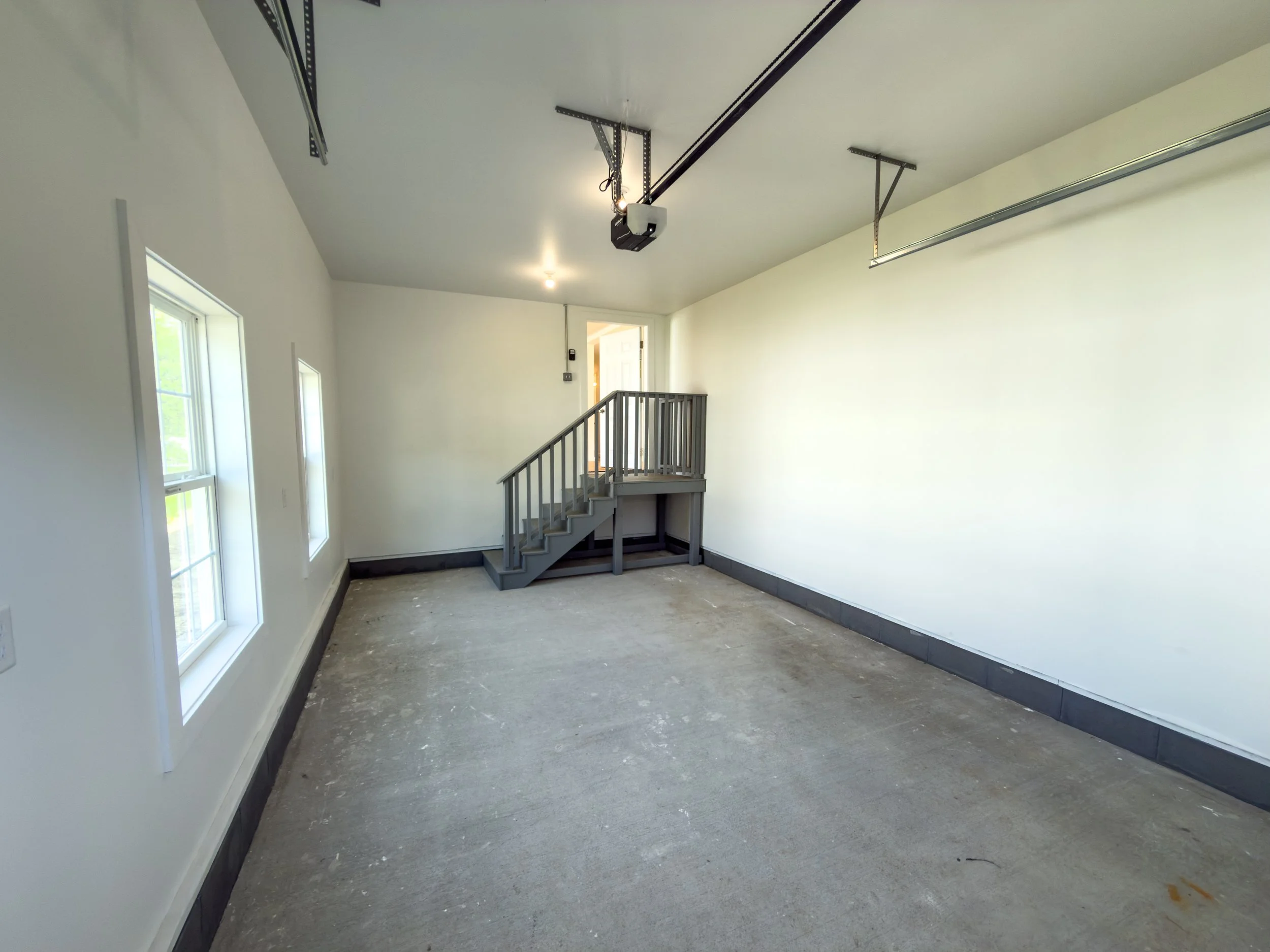 Interior of a garage with white walls, large side windows, a staircase with railings, and garage door tracks on the ceiling.