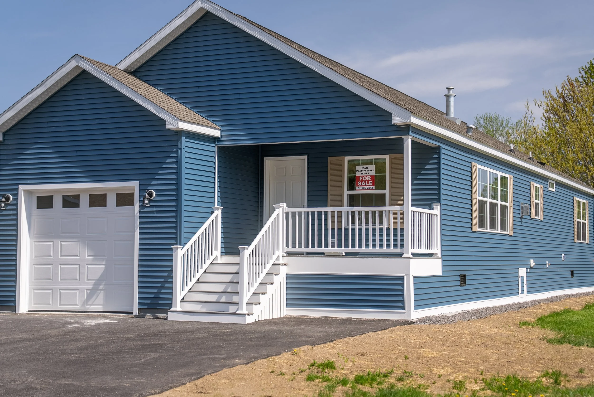 Blue manufactured home with white garage door and front porch, for sale sign in window, stairs leading to front door, green trees in background, partly cloudy sky.