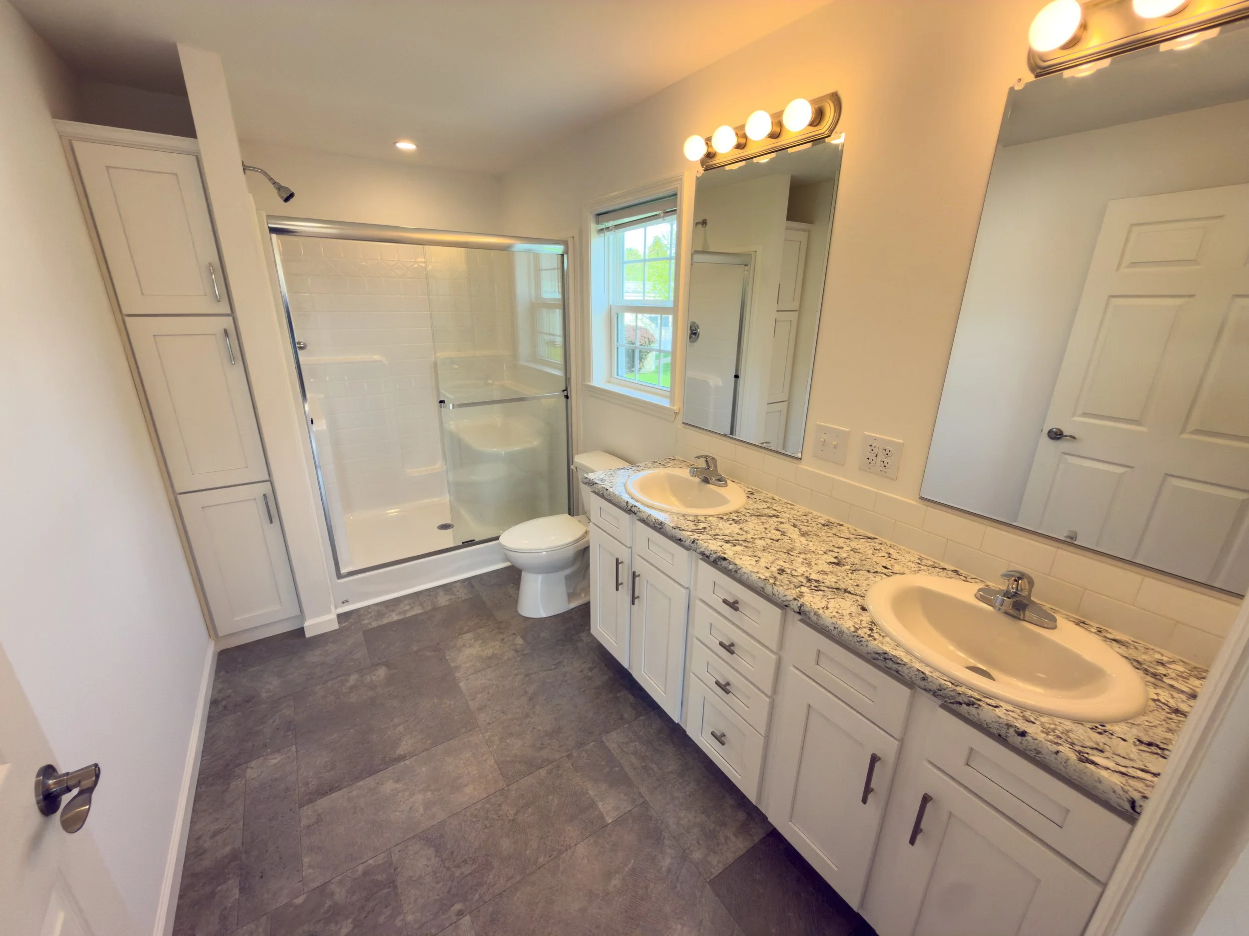 Elegant master bathroom with a double-sink vanity, large mirror, walk-in glass shower, toilet, tile flooring, and a window with views of greenery — a luxurious bathroom in a Manufactured Home
