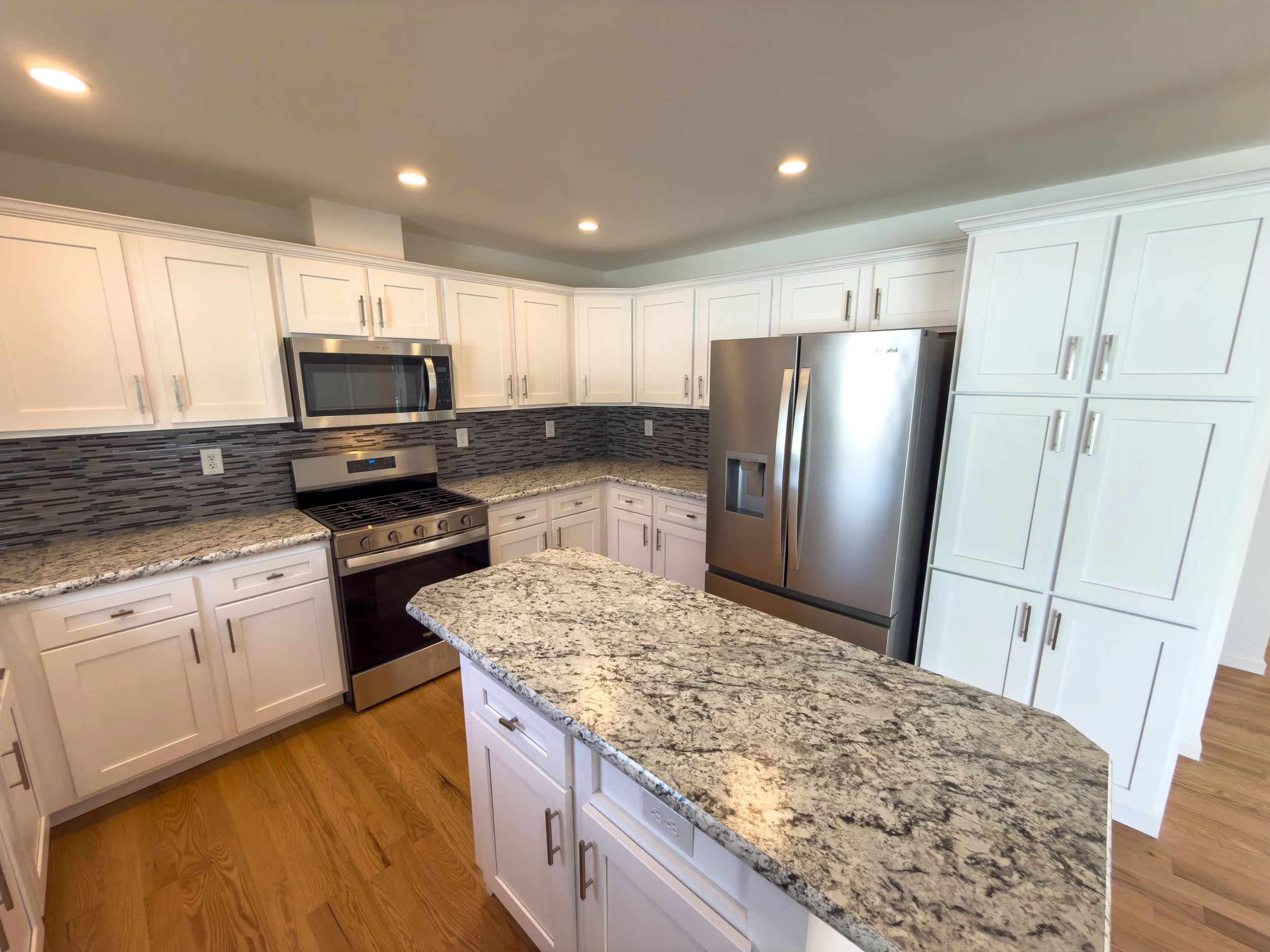 Well-equipped kitchen with white cabinets, countertop, stainless steel microwave, stove, and refrigerator, hardwood flooring, and a contemporary horizontal tile backsplash