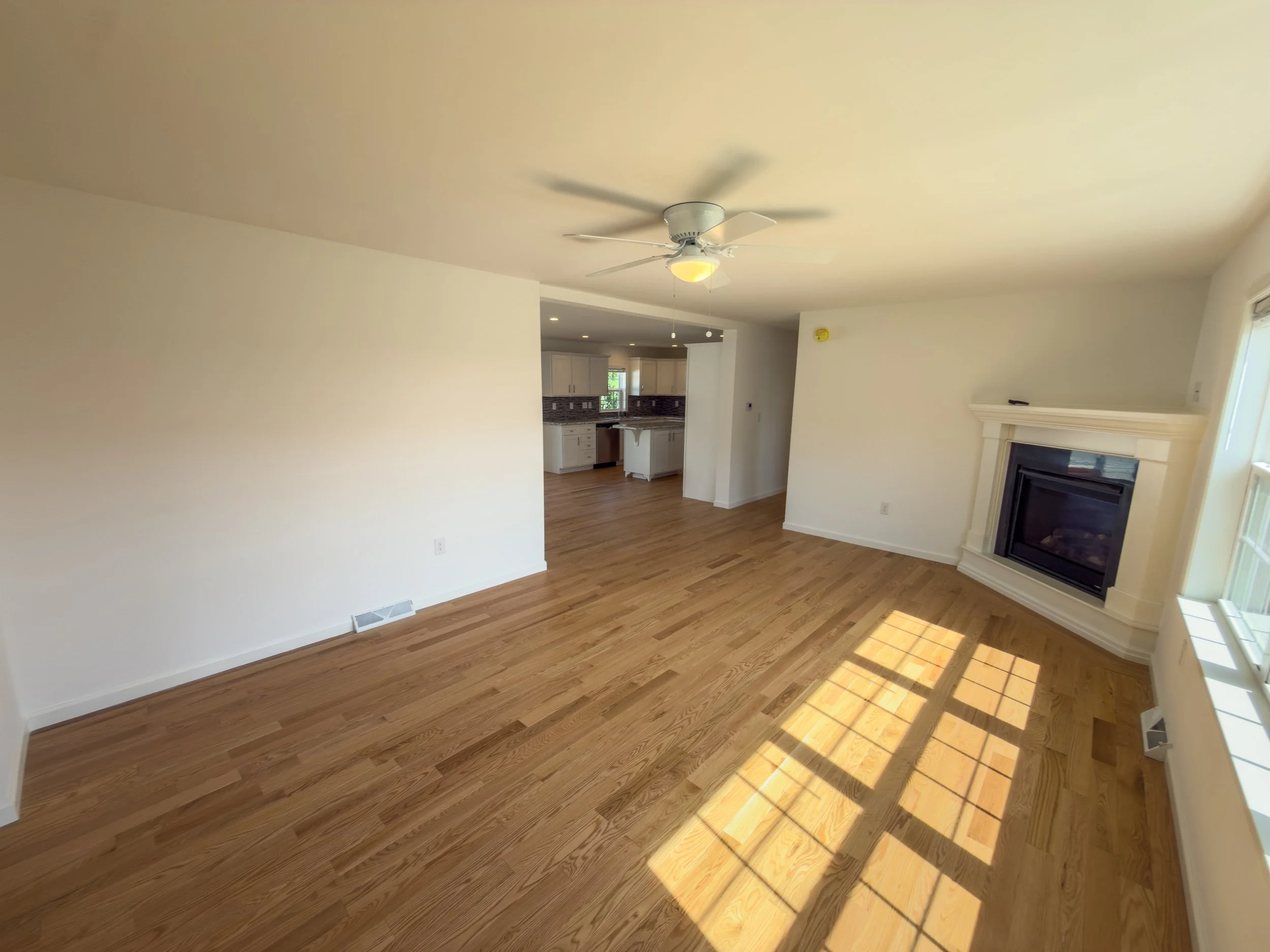 Sun-drenched open living room with warm hardwood flooring, a wood-burning fireplace, ceiling fan, and natural light streaming through windows — a welcoming interior in a Manufactured Home