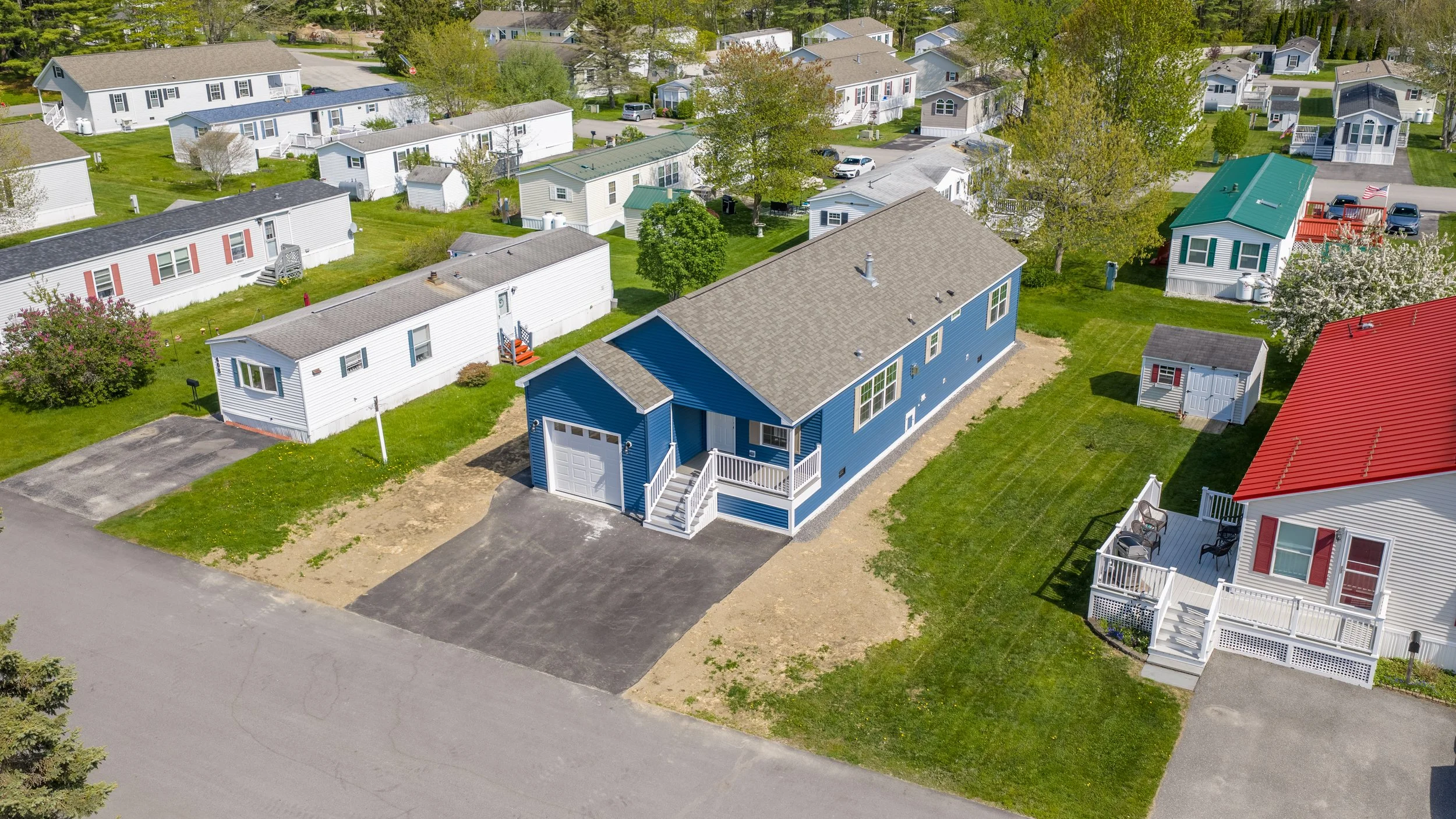 Aerial view of a residential neighborhood, Pinecrest, featuring a blue house with a front porch, driveway, and an adjacent white shed. Surrounding houses are visible with green lawns and trees.