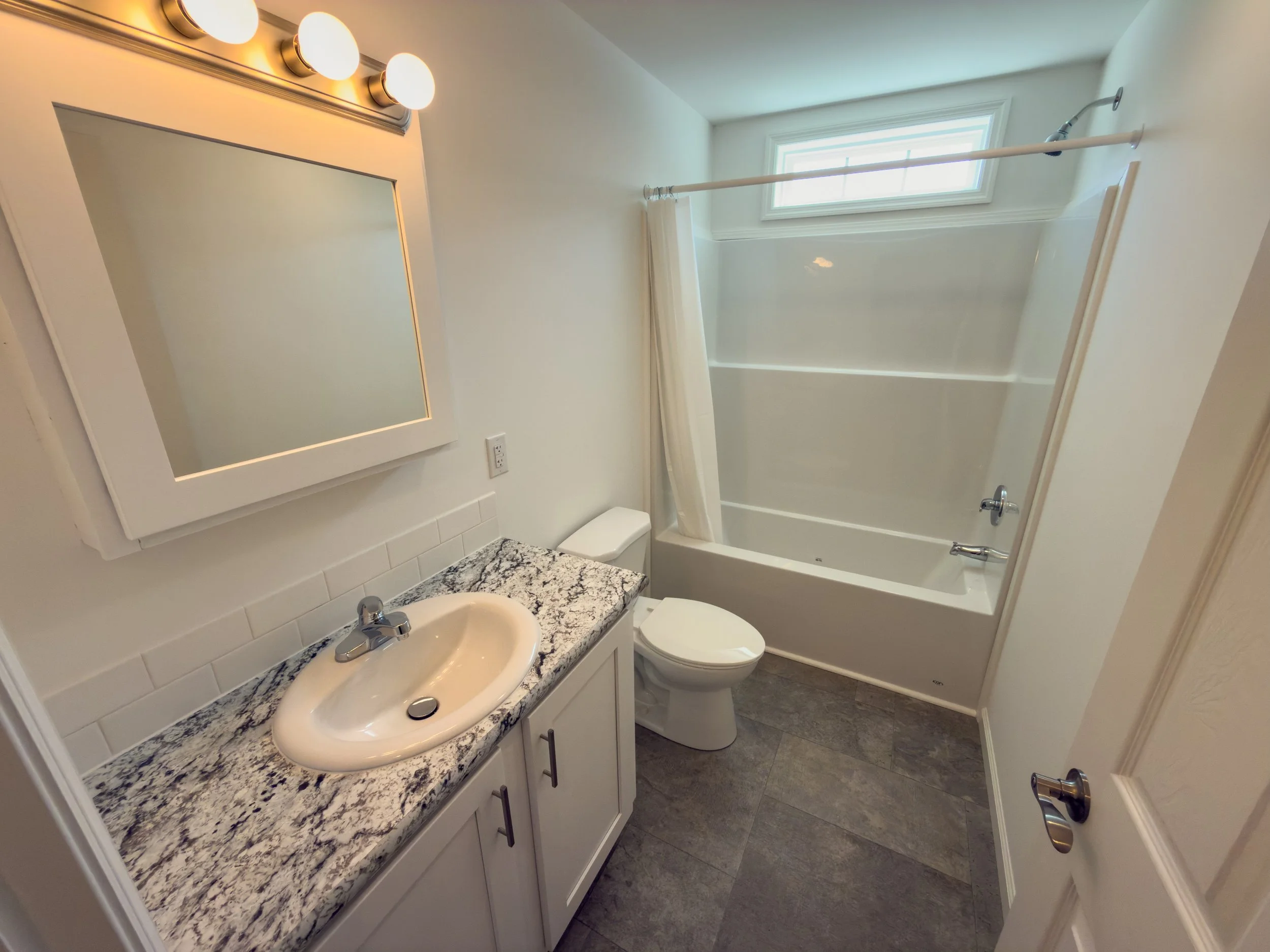 Clean full bathroom with a countertop vanity, white sink, lighted mirror, toilet, and shower-tub combination with window above — a well-appointed bathroom at State Manufactured Homes