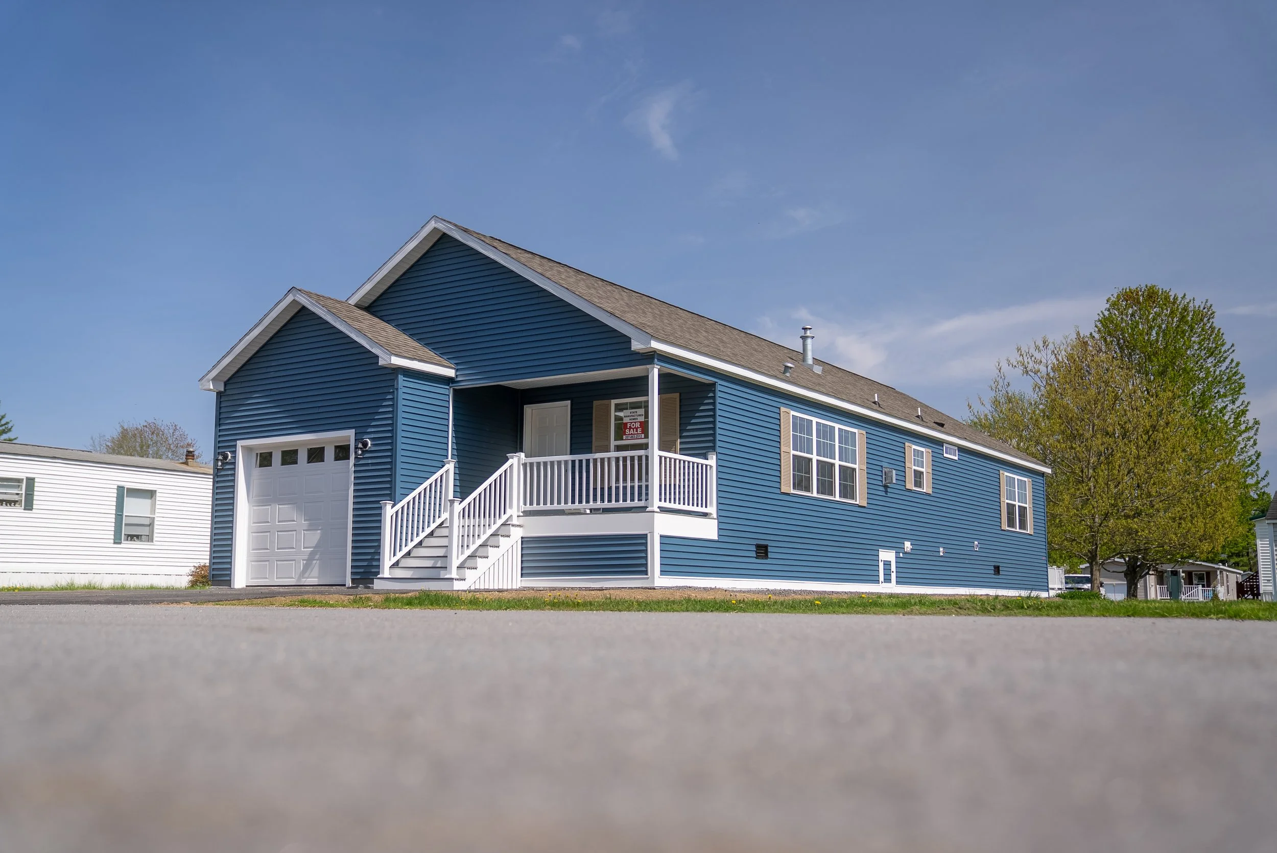 Blue house with front porch and stairs, white garage door, on a residential street with trees and a clear blue sky.