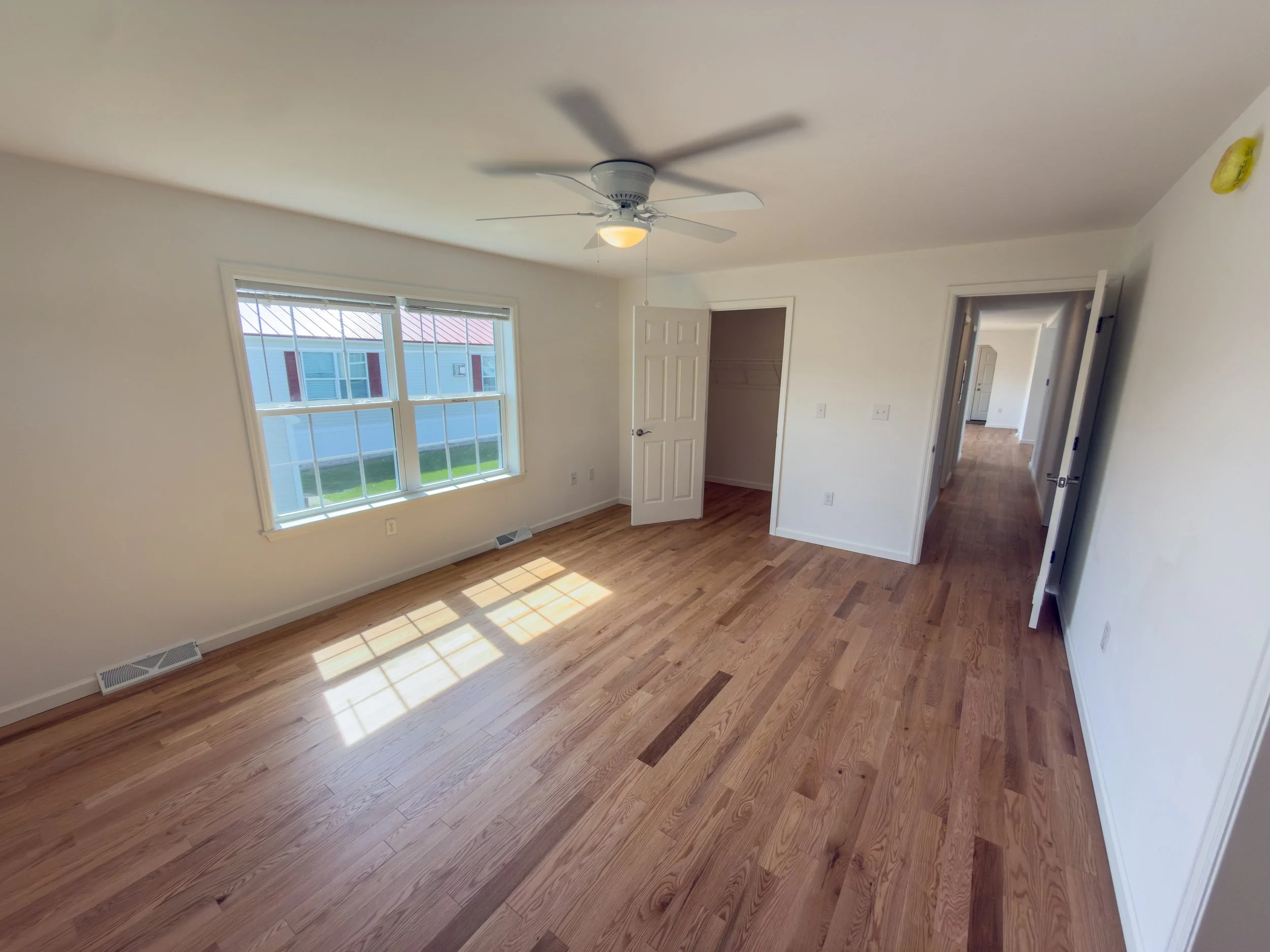 Spacious bedroom with hardwood floors, a ceiling fan, large bright window, open closet, and a hallway entry — a well-sized bedroom ready to personalize in a Manufactured Home