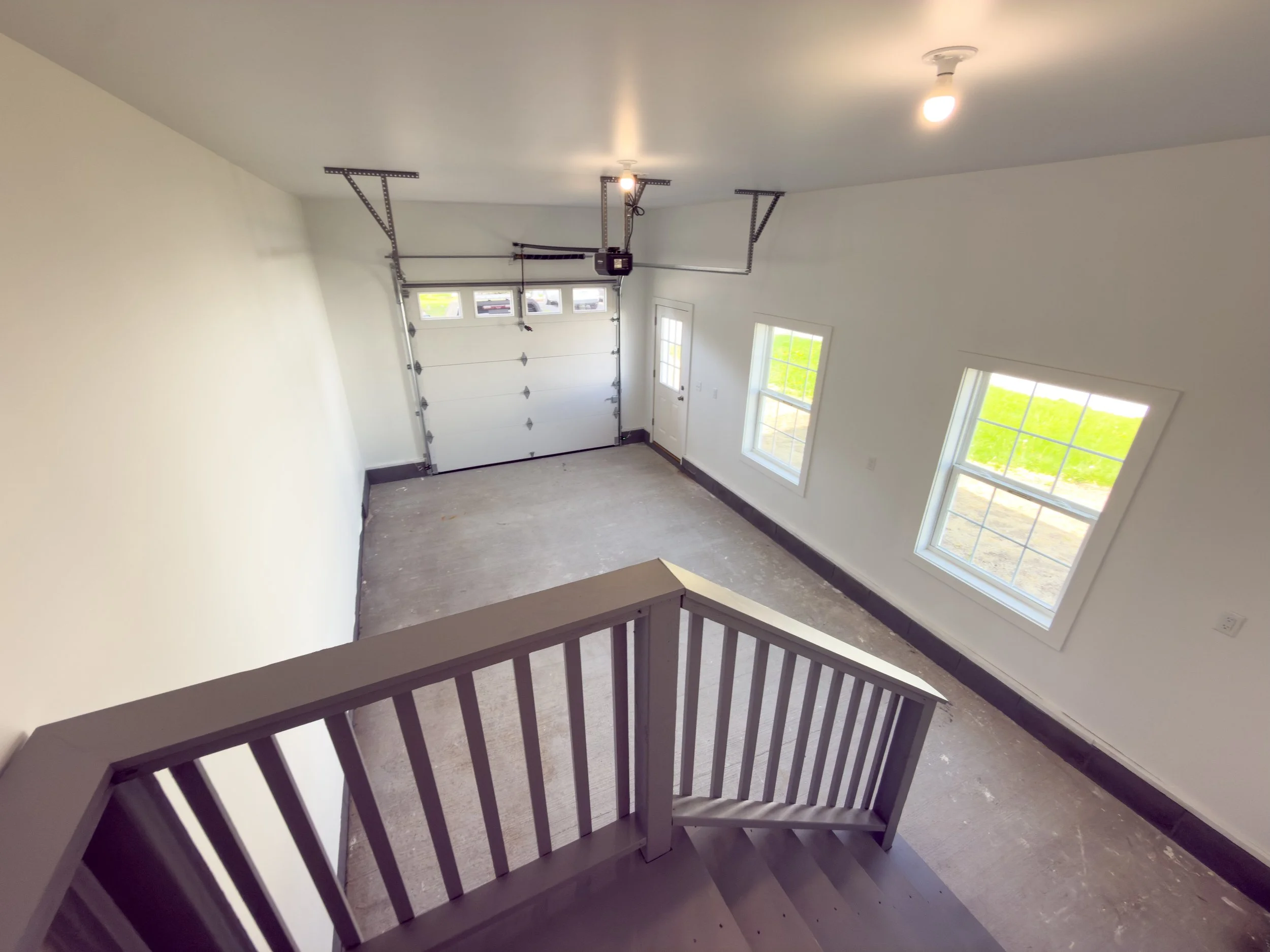 Empty garage with a white garage door, windows, and a door leading outside. It is viewed from the top of a staircase with a wooden railing.