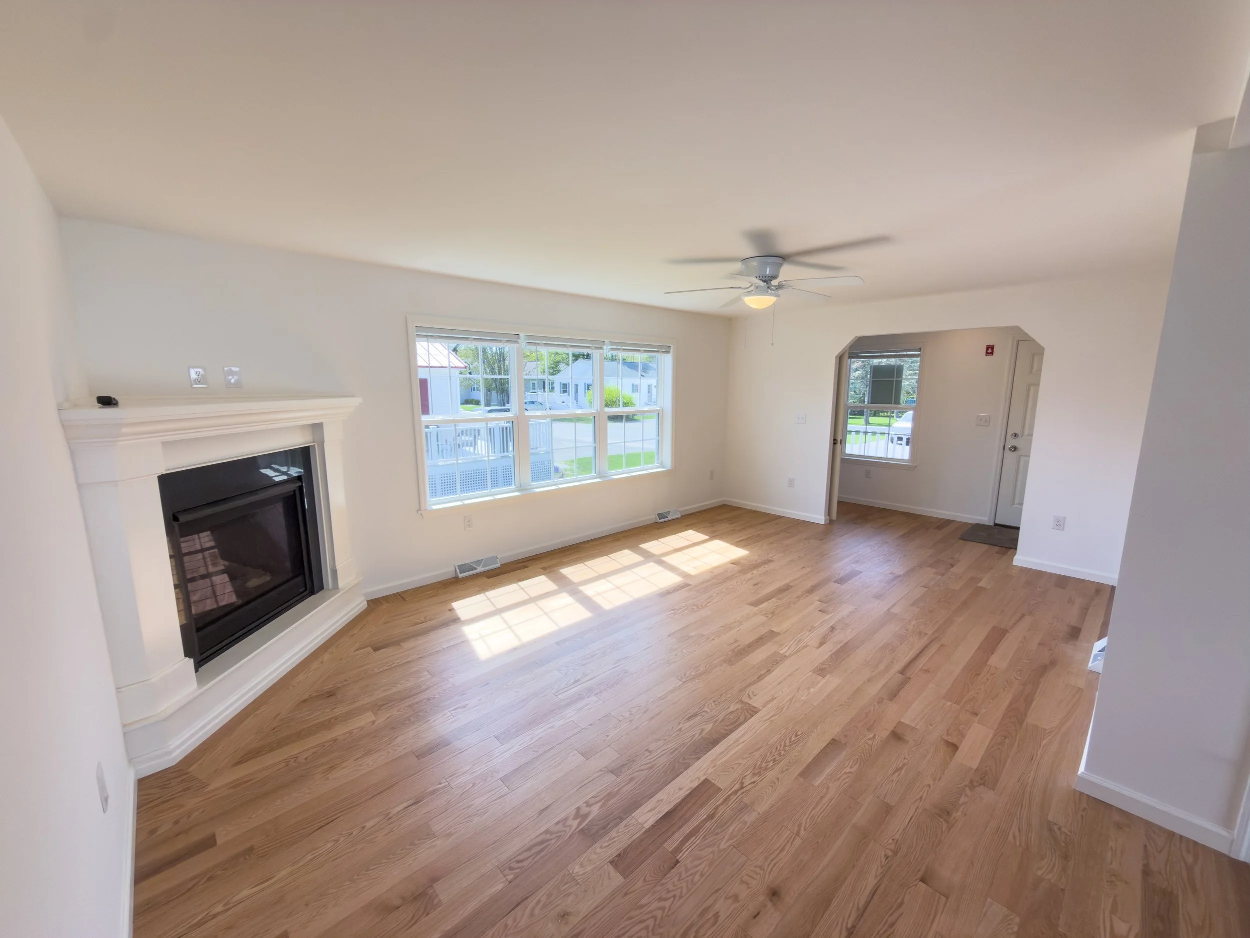 Spacious living room with gleaming hardwood floors, a fireplace, ceiling fan, and a large picture window flooding the space with natural light — an inviting main living area in a Manufactured Home