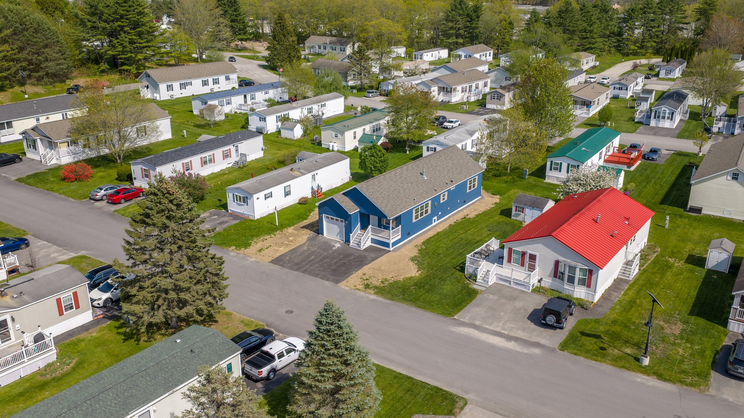 Wide aerial view of a vibrant manufactured home community with diverse single-story homes, lush green lawns, mature trees, and paved streets — a welcoming neighborhood setting offered by State Manufactured Homes