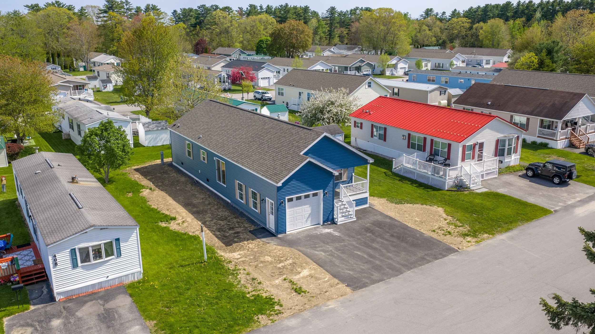 Elevated aerial view of a blue manufactured home with attached garage among neighboring homes, lush trees, and paved driveways in a well-established community by State Manufactured Homes