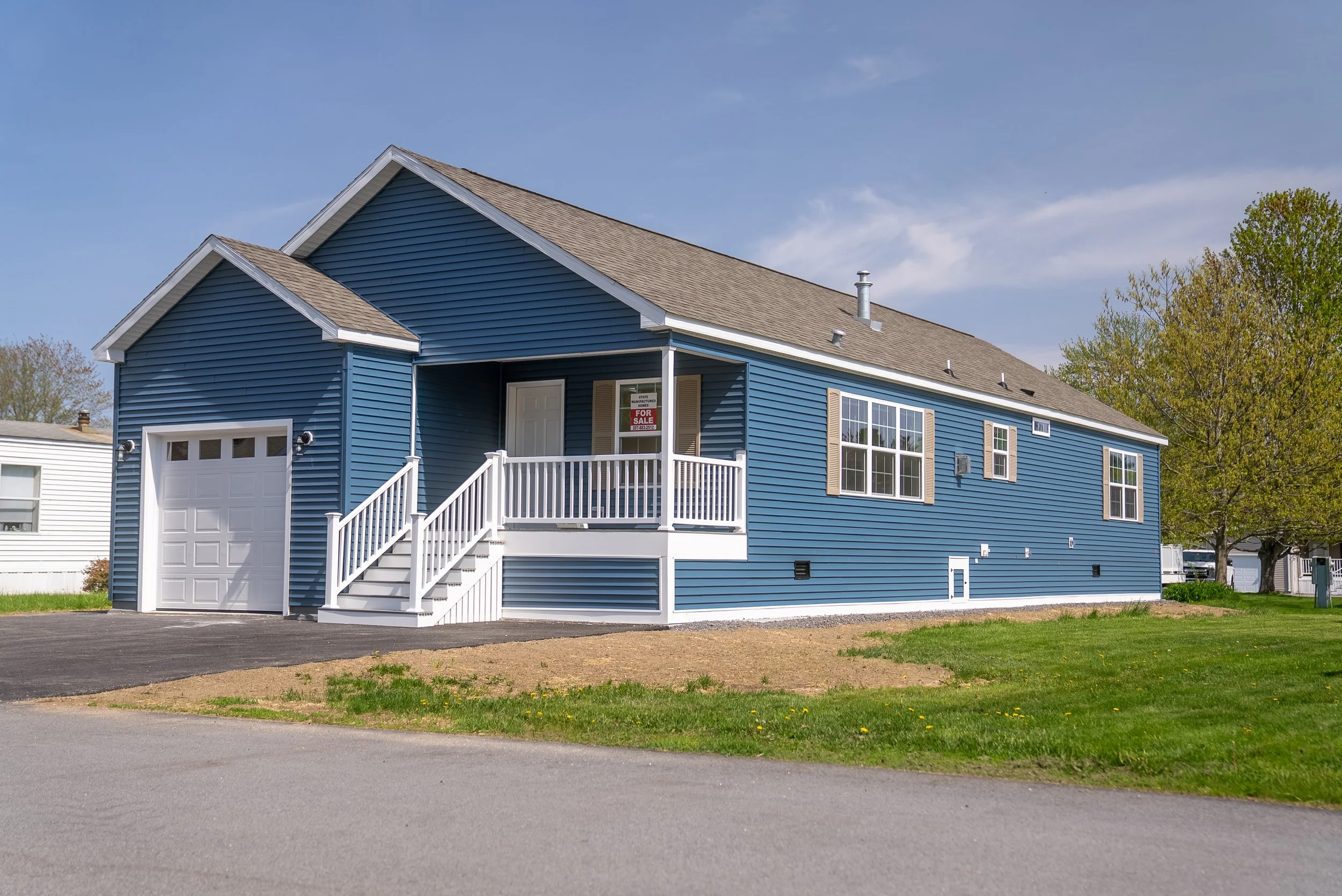 A blue house with white trim, a white garage door, a front porch, and a staircase leading up to the front door. There is a 'For Sale' sign in the front window, and the house is surrounded by a grassy yard with trees in the background under a clear blue sky.