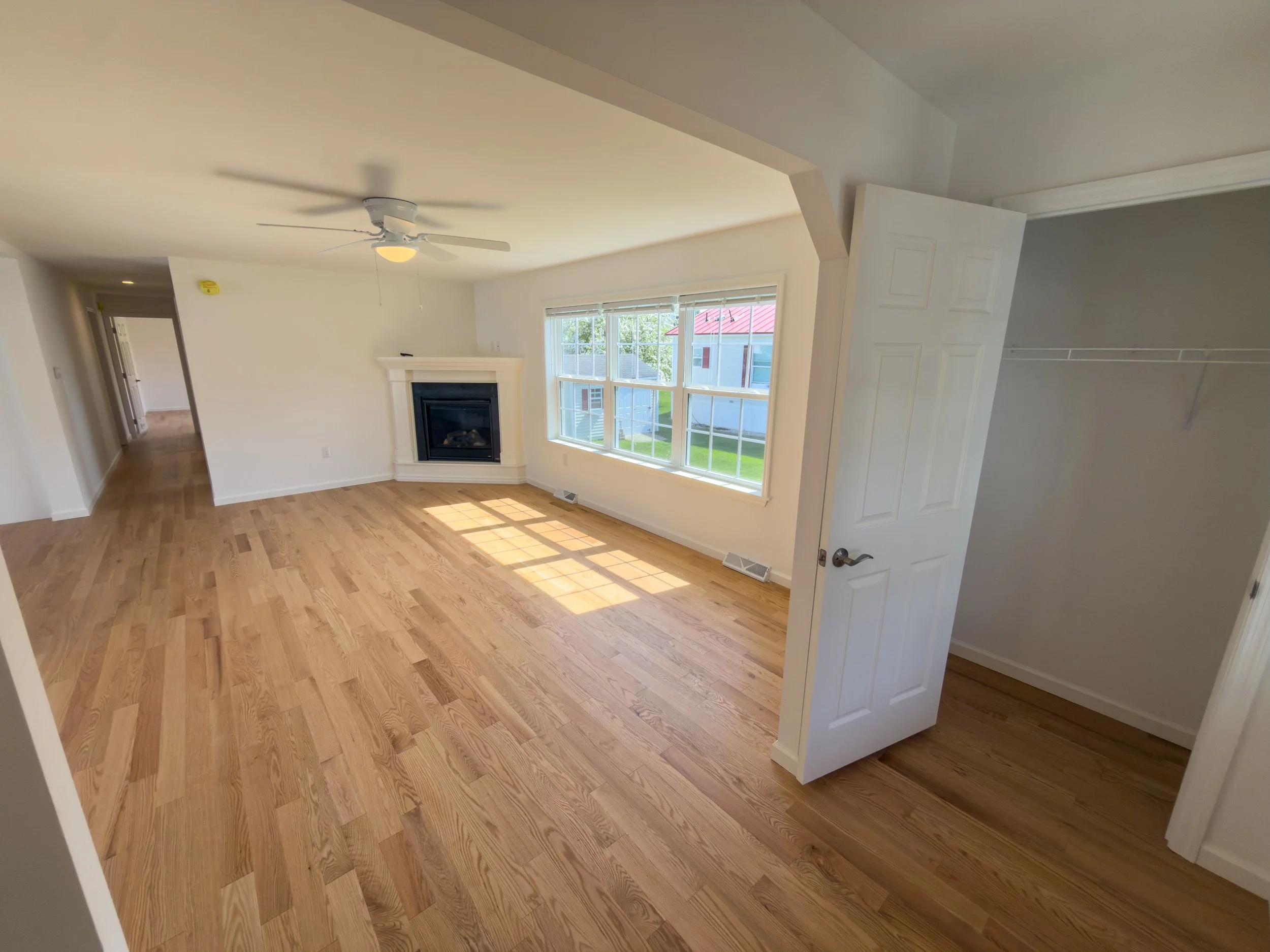 Bright and open living room with gleaming hardwood floors, white walls, large windows for natural light, a ceiling fan, and a fireplace — move-in ready interior space in a new manufactured home