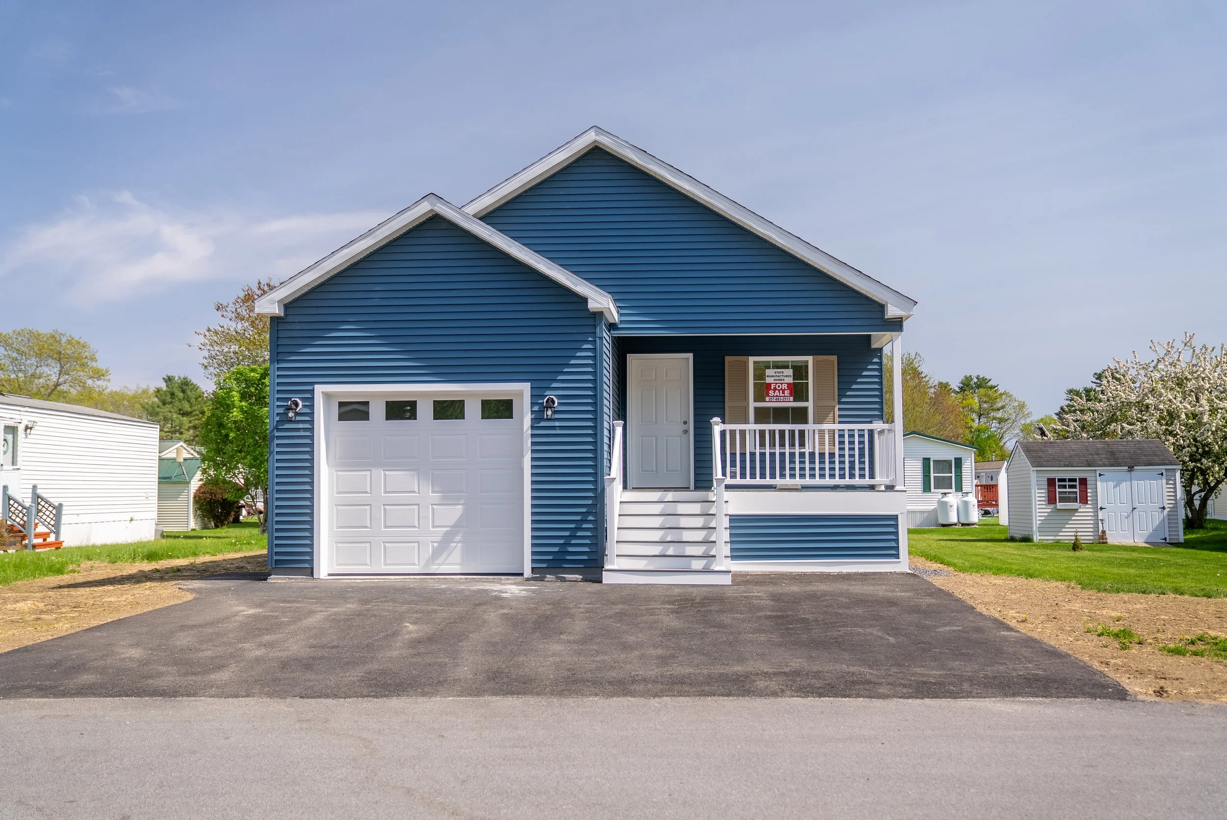 Blue house with white garage door and front stairs, for sale sign in front window, green grass, neighboring houses, trees, and clear sky in the Pinecrest Community.