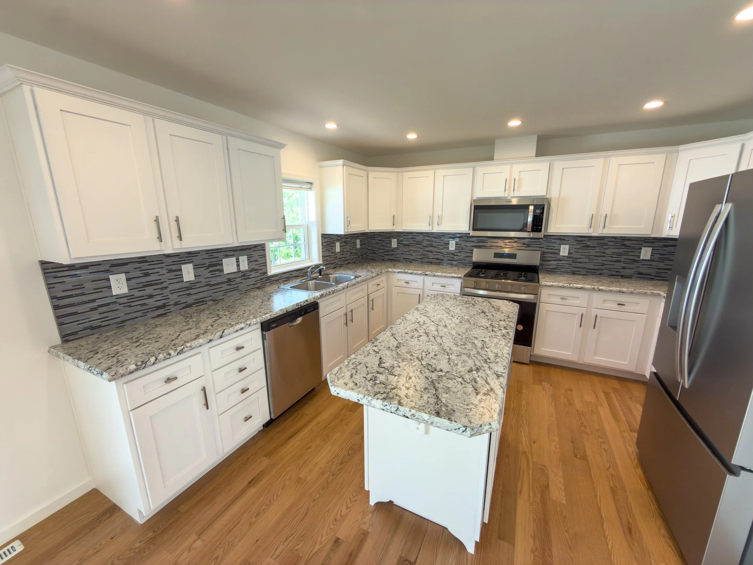A modern kitchen with white cabinets, countertops, stainless steel appliances including a microwave and a refrigerator, and a window letting in natural light.