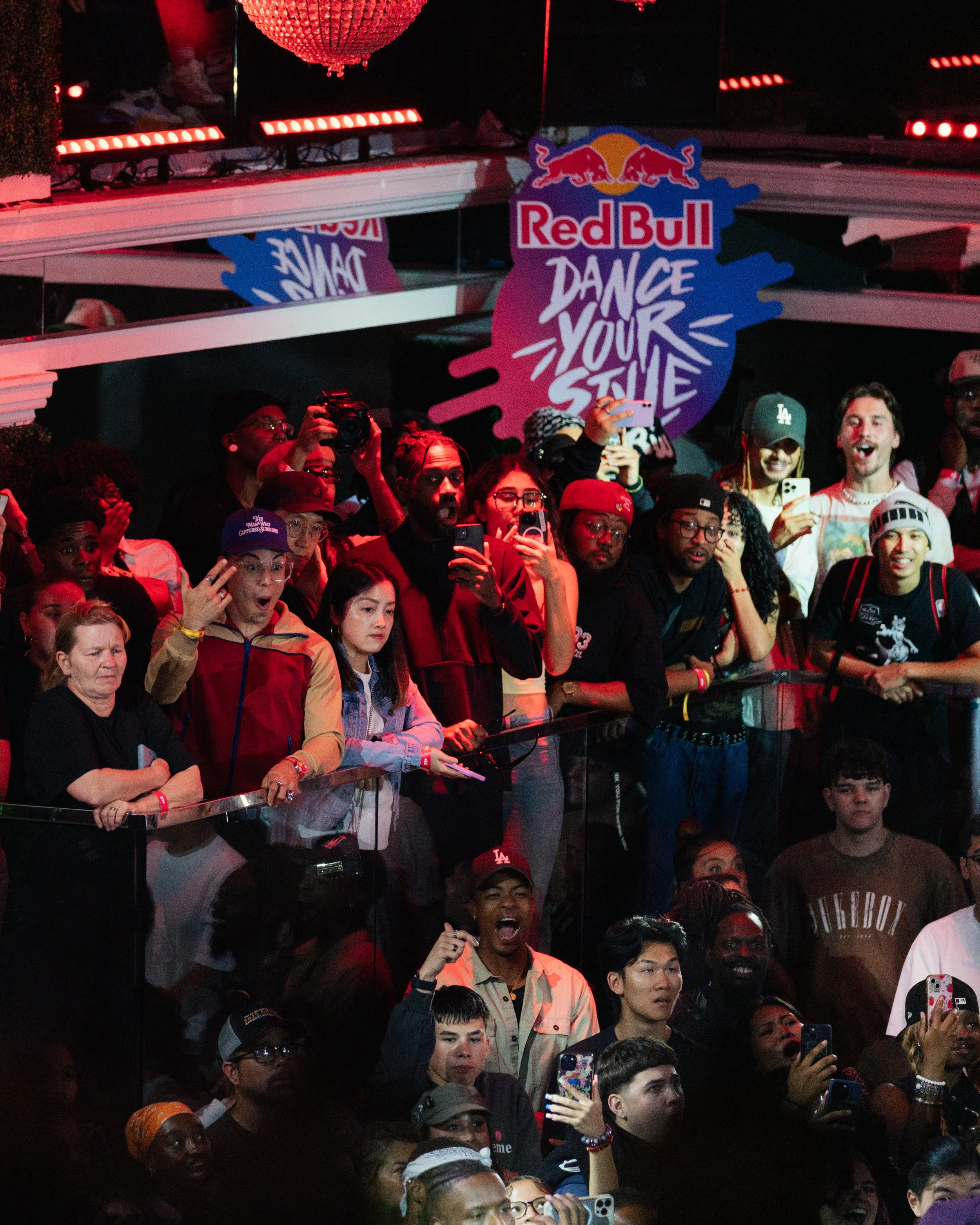 Crowd of people at a dance event under a sign that says 'Red Bull Dance Your Style' illuminated with red and pink lights.