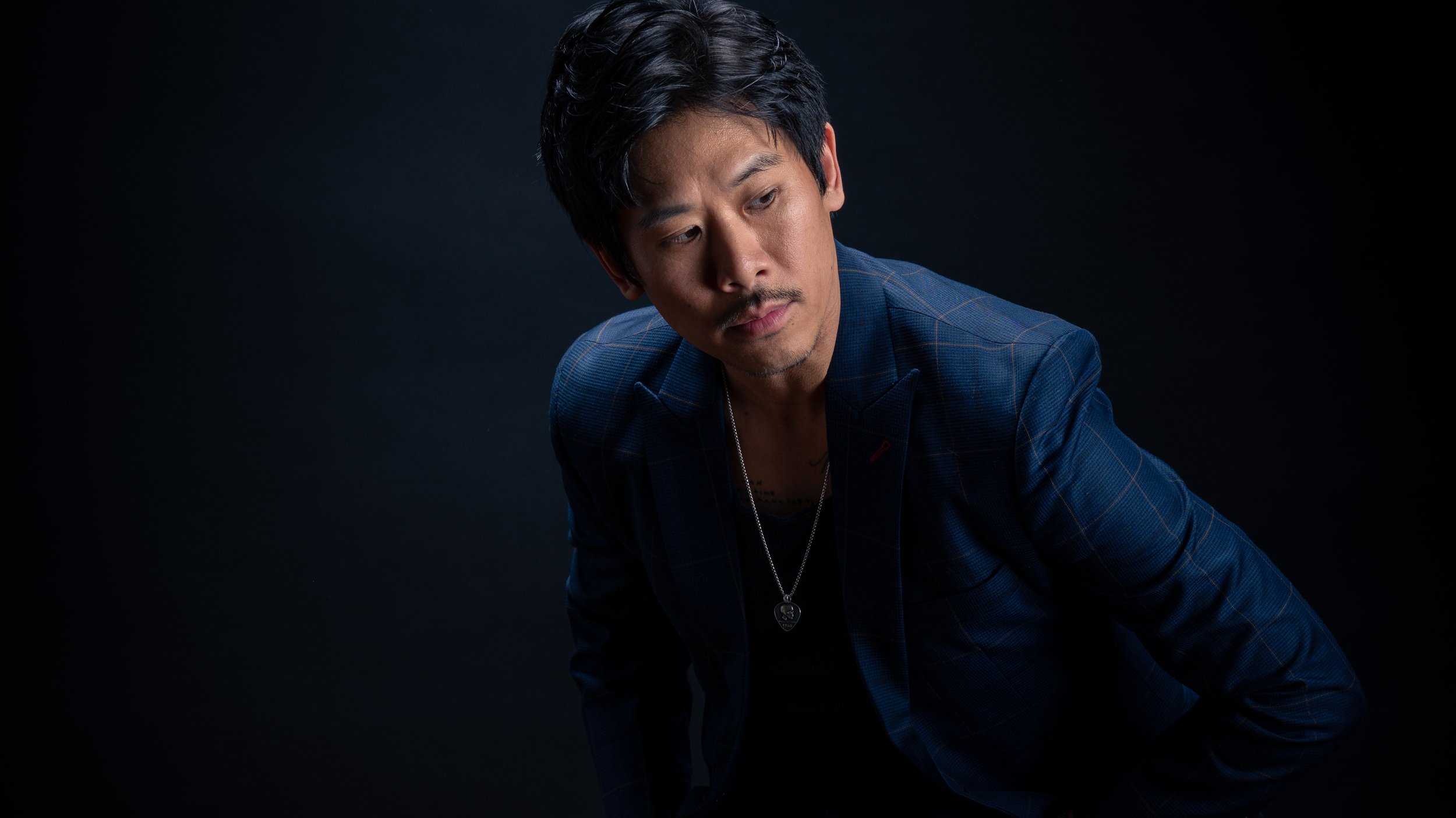 A man with dark hair and a mustache in a blue checkered blazer, looking to the side against a dark background.