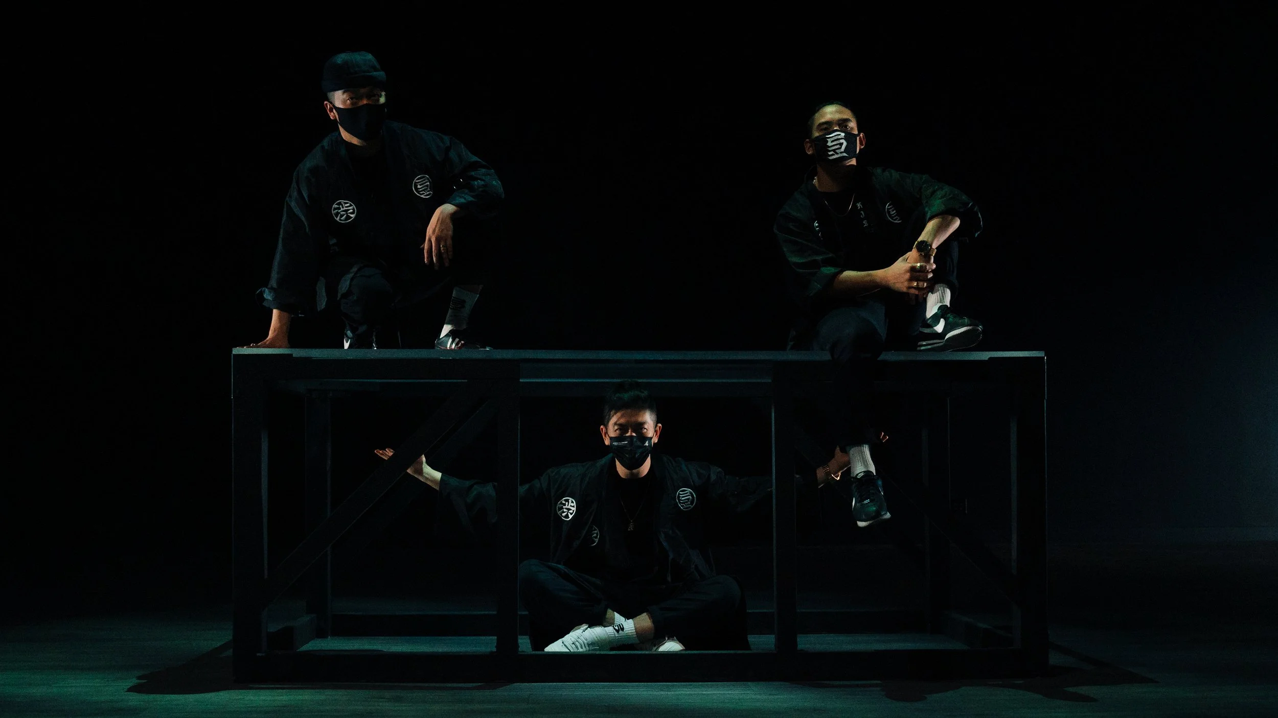 Three individuals wearing black masks and black clothing pose on and under a black table in a dark space, with a spotlight highlighting their presence.
