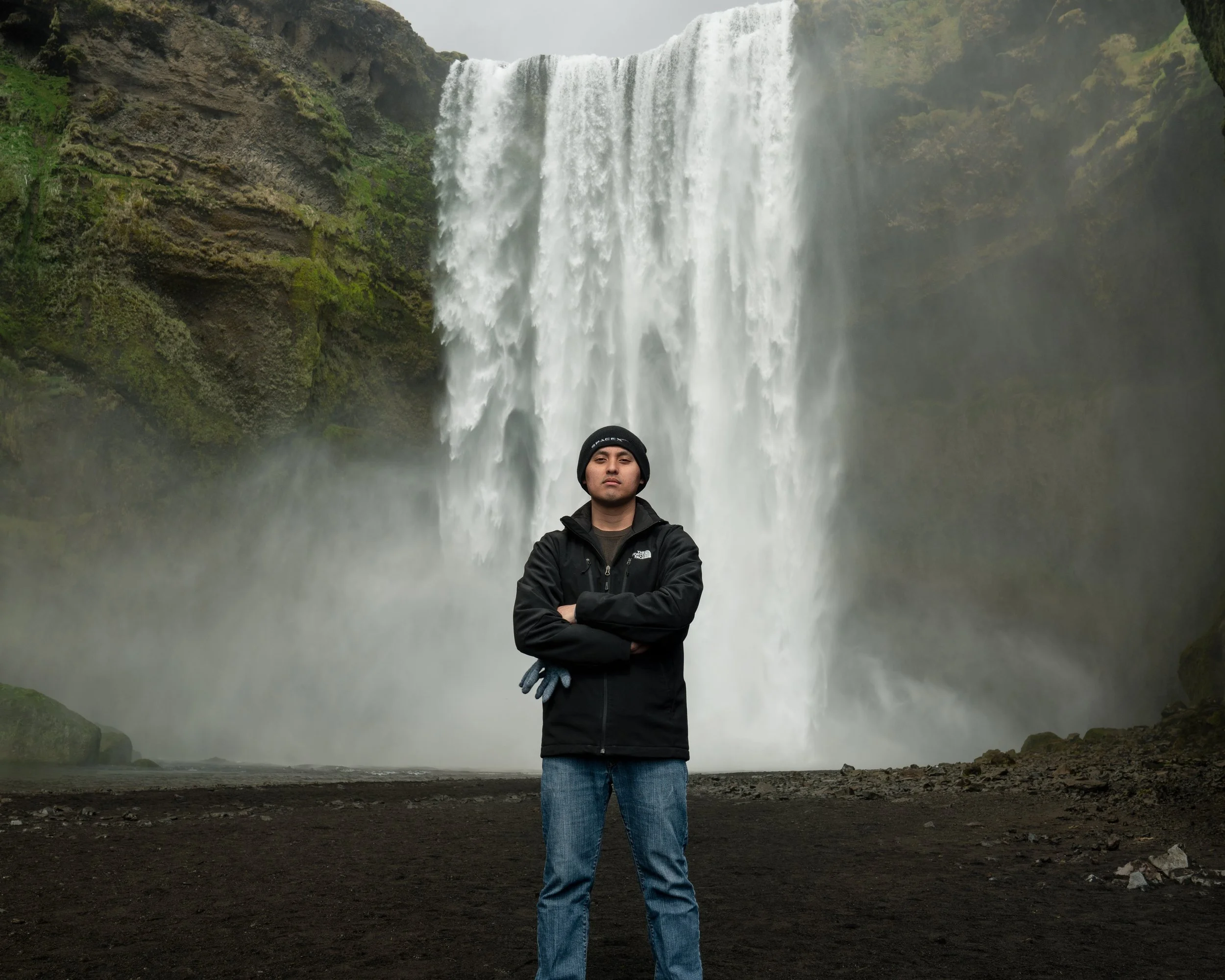 A man in a black jacket and beanie stands with crossed arms in front of a large waterfall surrounded by green moss-covered rocks.