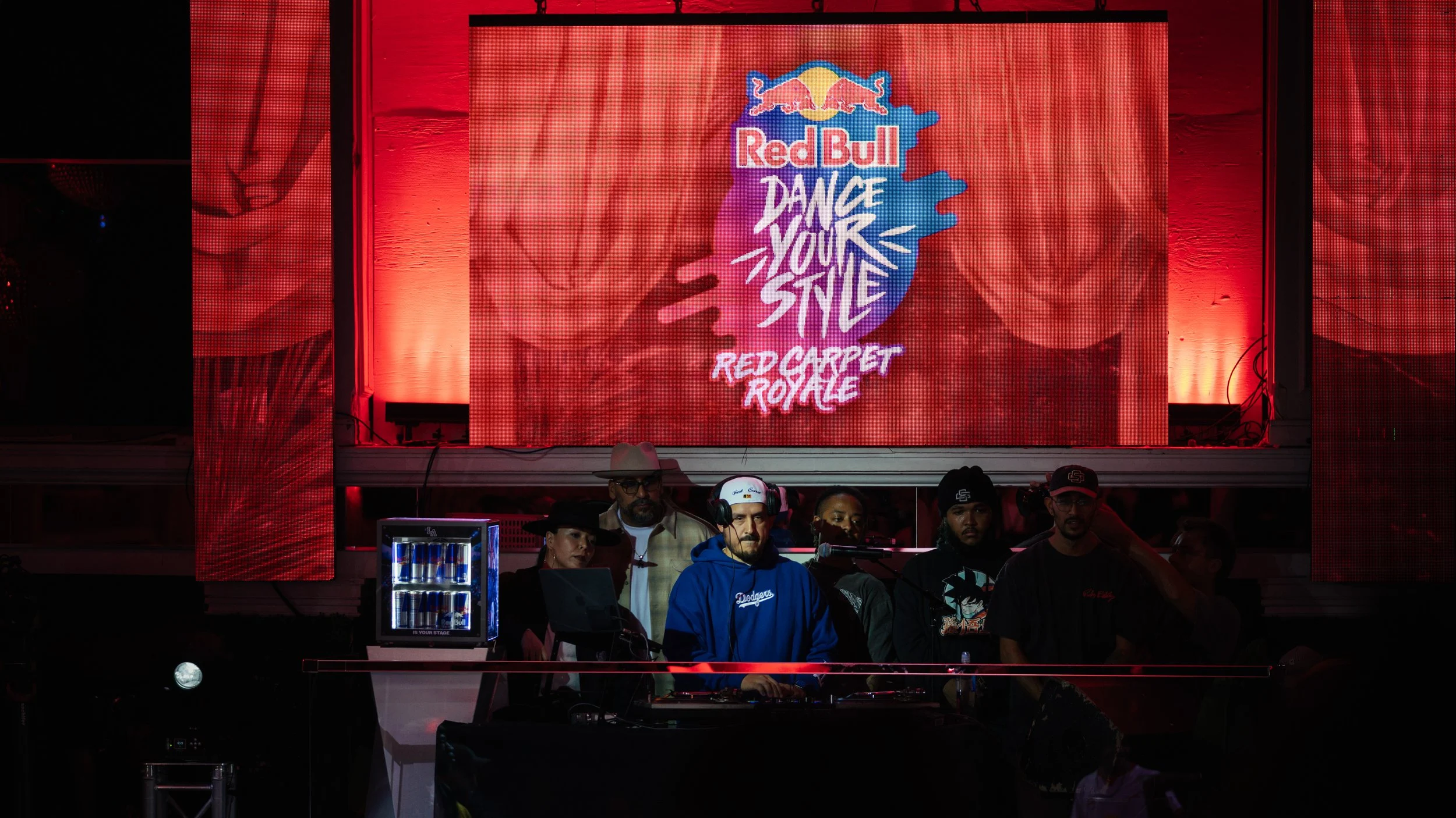 A DJ performs at the Red Bull Dance Your Style event on a stage with a large digital screen displaying the event logo and name, featuring red and pink curtains in the background and people standing behind the DJ.