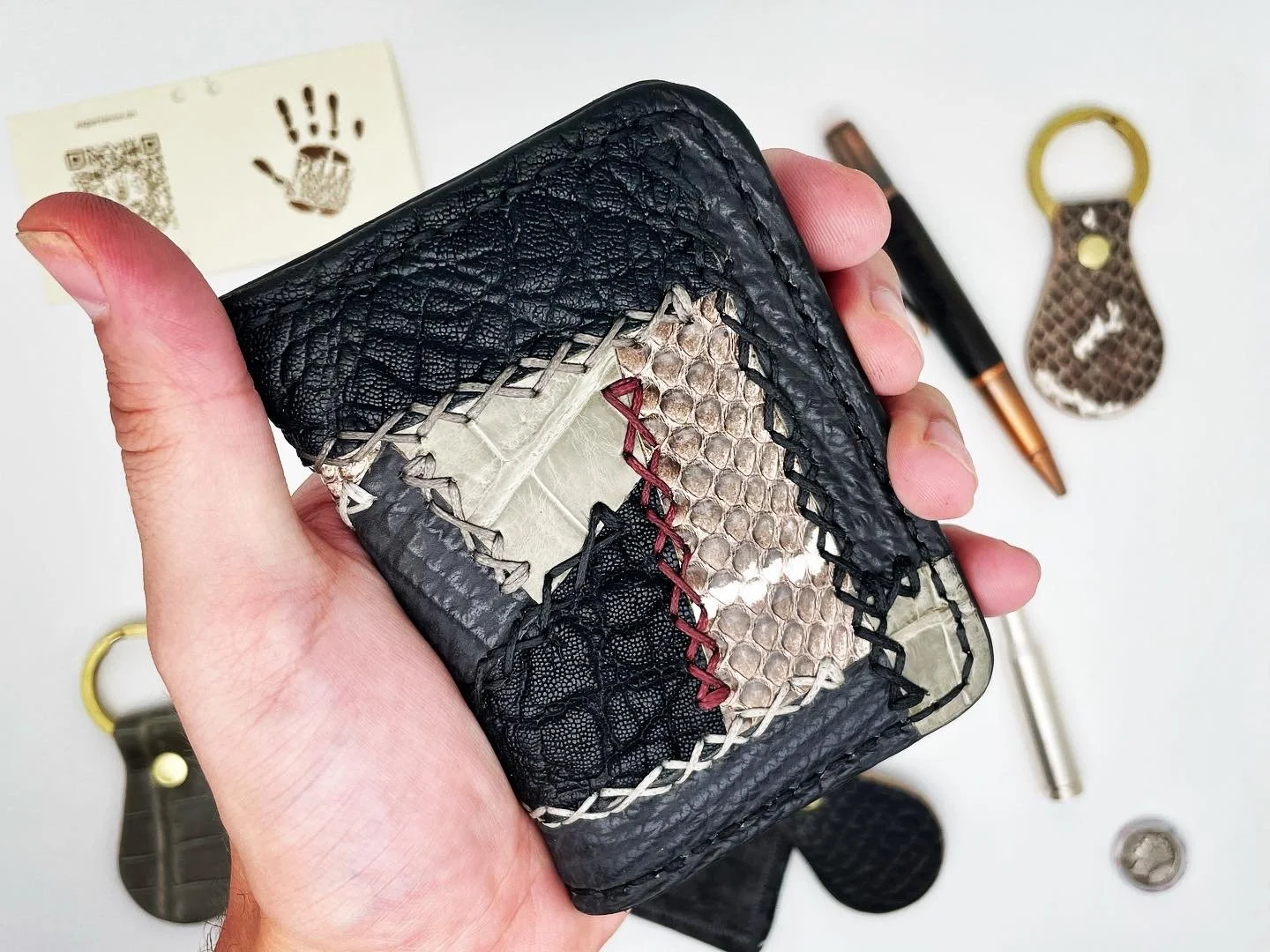 A hand holding a small black wallet with snake skin pattern and cross-stitch embroidery, surrounded by various small leather items, a pen, a bottle opener, and a coin on a white surface.